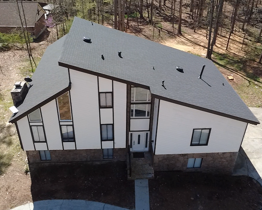 A two-story house with a white exterior, black trim, a gray roof, and multiple windows, situated in a wooded area.