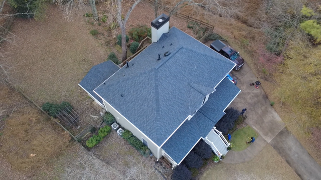 Aerial view of a house with a dark gray roof and a chimney, surrounded by trees, a driveway, and some outdoor equipment.
