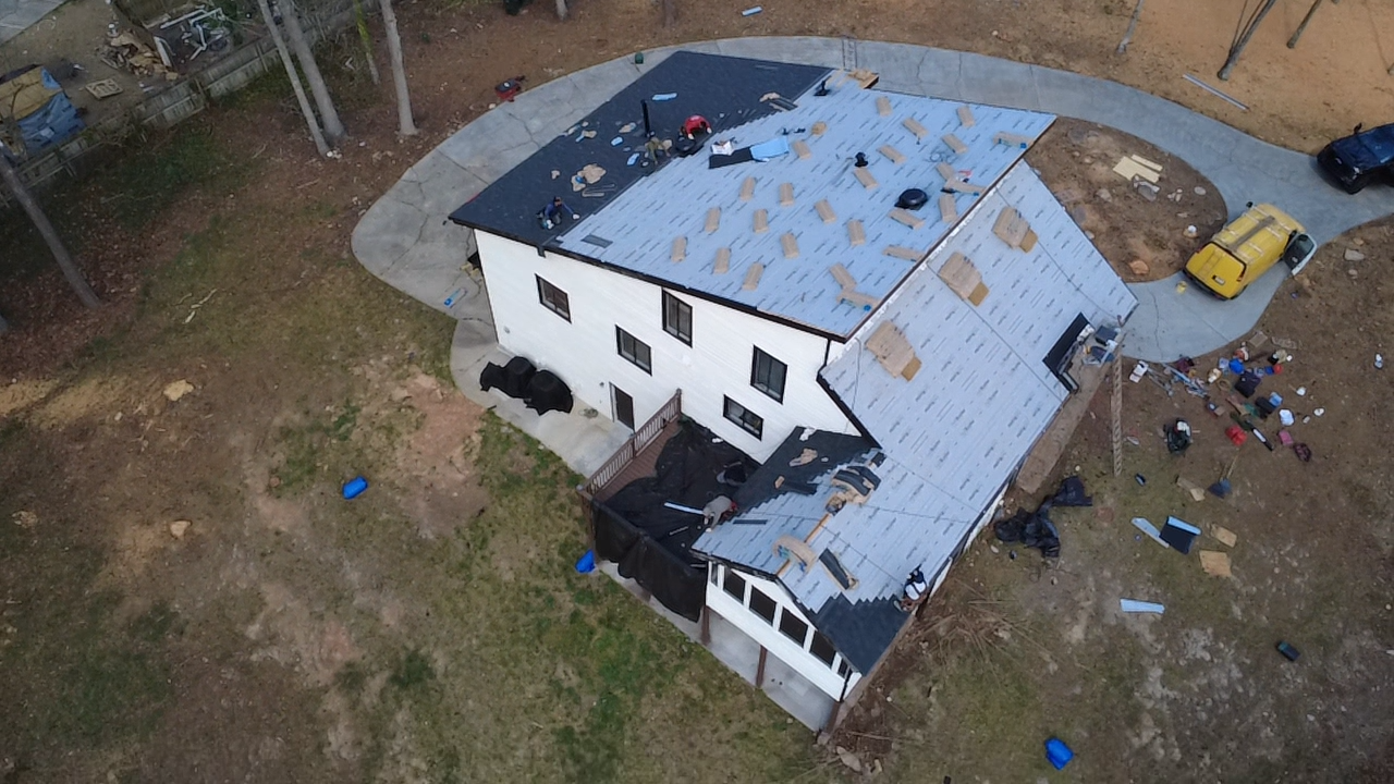 A house under construction or renovation with roofing work ongoing, surrounded by construction debris and vehicles, on a dirt lot with some grass and trees nearby.