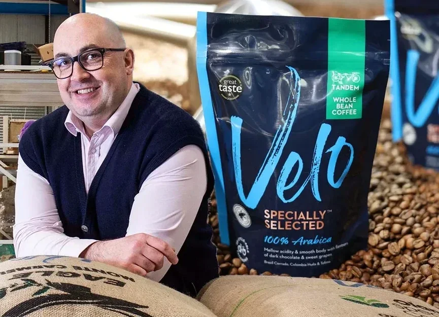 A smiling man with glasses and a bald head leaning on a table with burlap bags of coffee beans, next to a large stand-up pouch of Velo coffee labeled 'Specially Selected 100% Arabica' with a background of coffee beans.