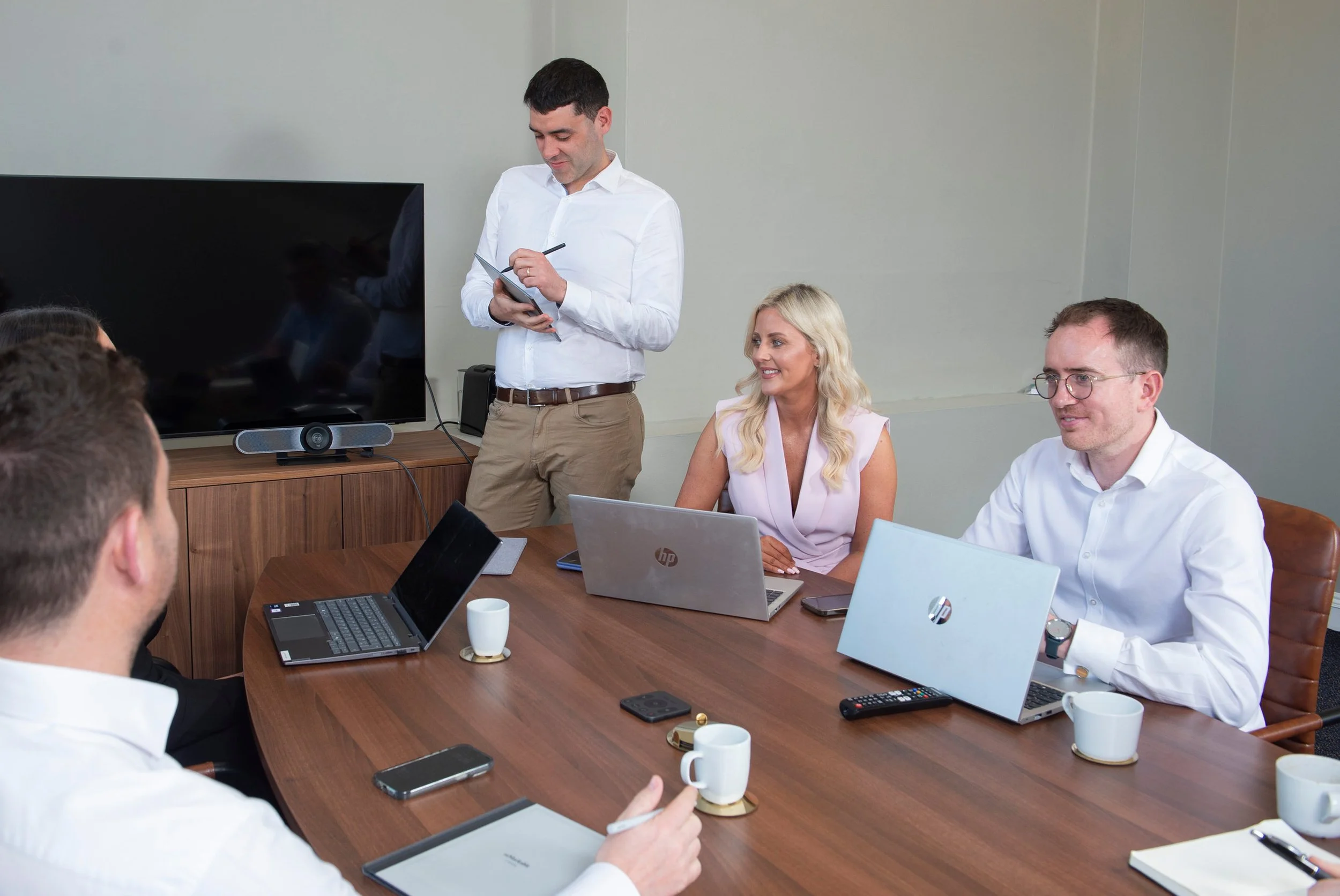 Business meeting with four people seated around a wooden table in a conference room. One man is standing, writing on a notepad, while three others listen and look at laptops or tablets.