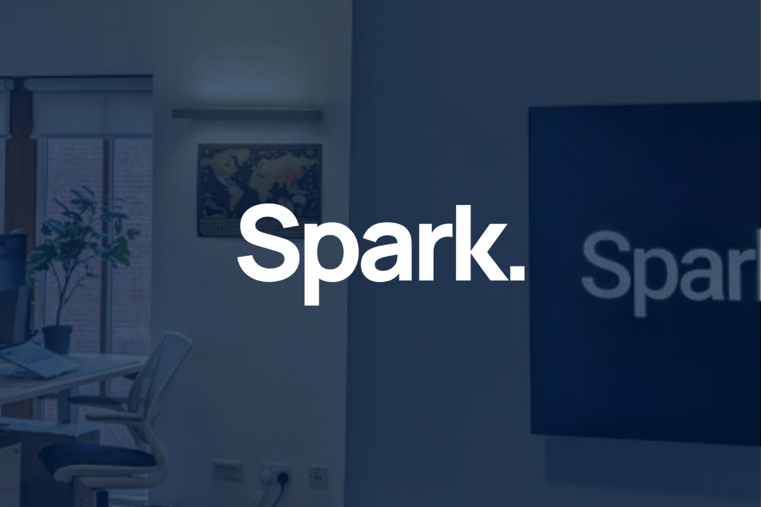 Office interior with a desk, chair, computer, indoor plant, world map on the wall, and 'Spark.' logo on a screen.