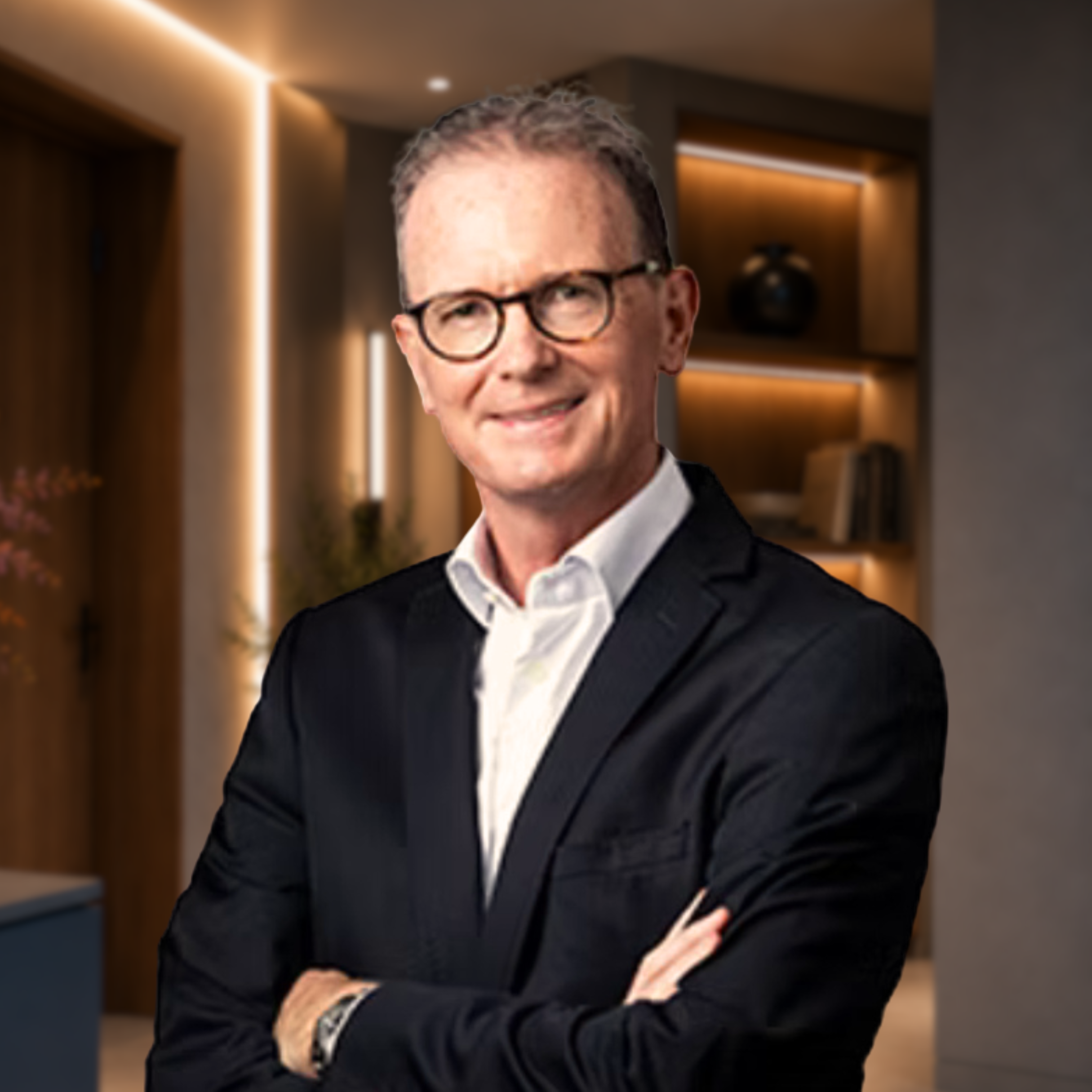 A man with glasses and a suit standing in a modern, warmly lit room with wooden decor and shelves, smiling at the camera.