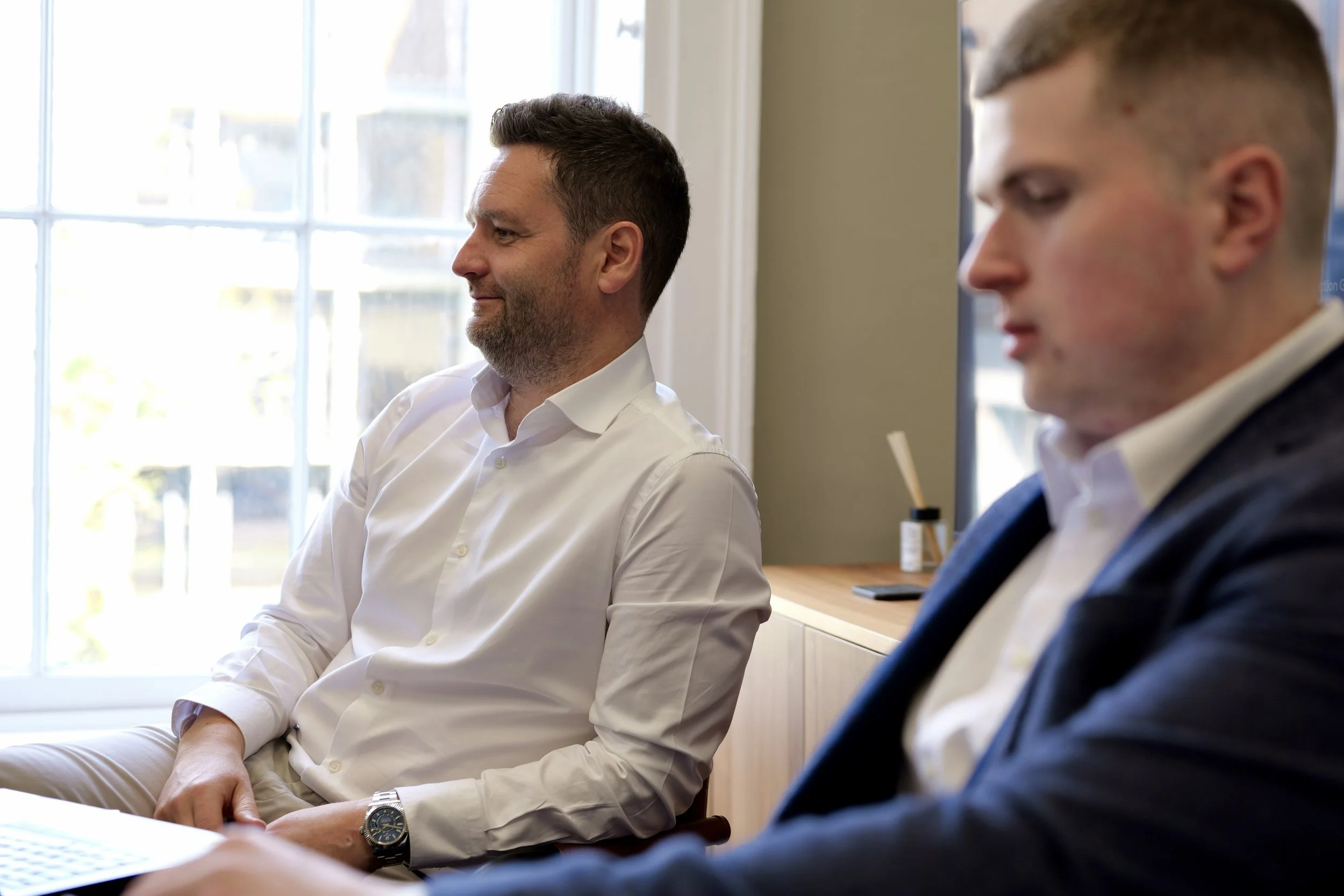 Two men sitting in a meeting or classroom, with one man in the foreground and another man in the background near a window, both dressed in business casual attire.