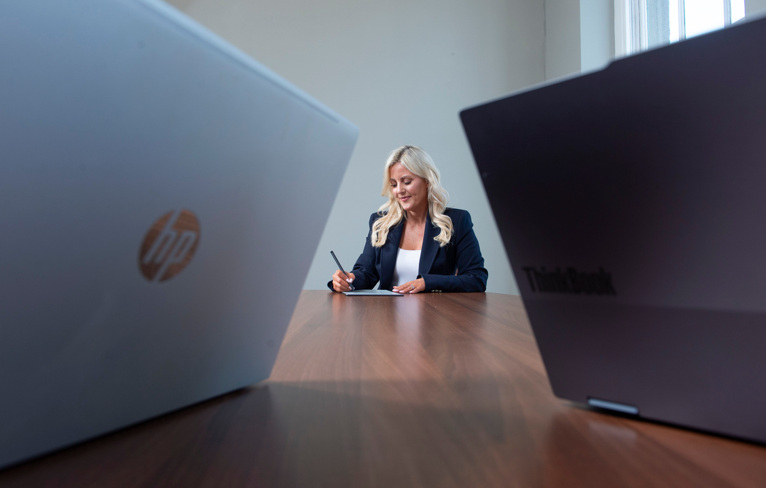 A woman with blonde hair sitting at a wooden table, writing on a notepad, with two laptops in the foreground, one white HP and one black Lenovo, in a bright room with large windows.