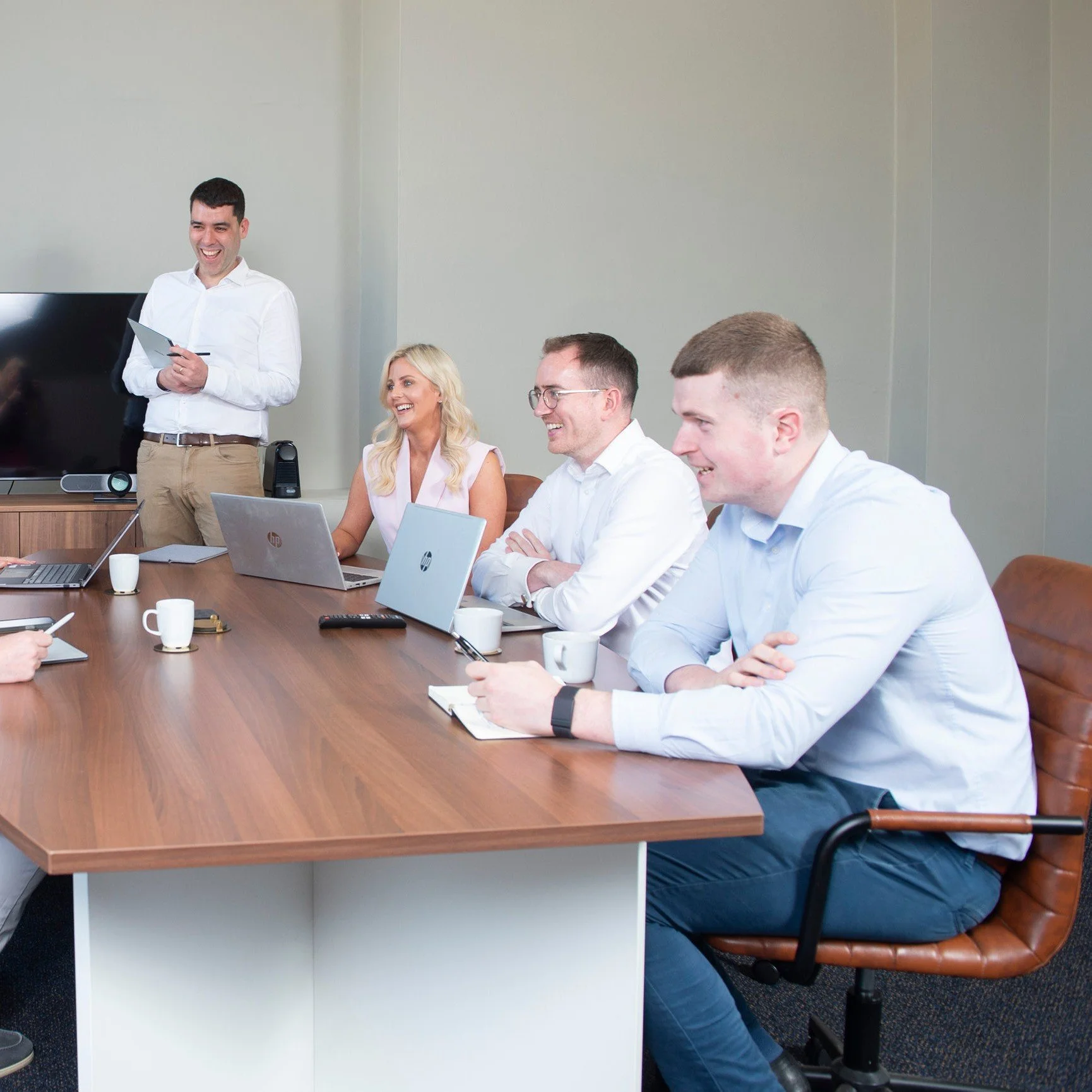 People sitting at a conference table with laptops and coffee mugs, engaged in a meeting, while a man stands laughing holding a tablet.