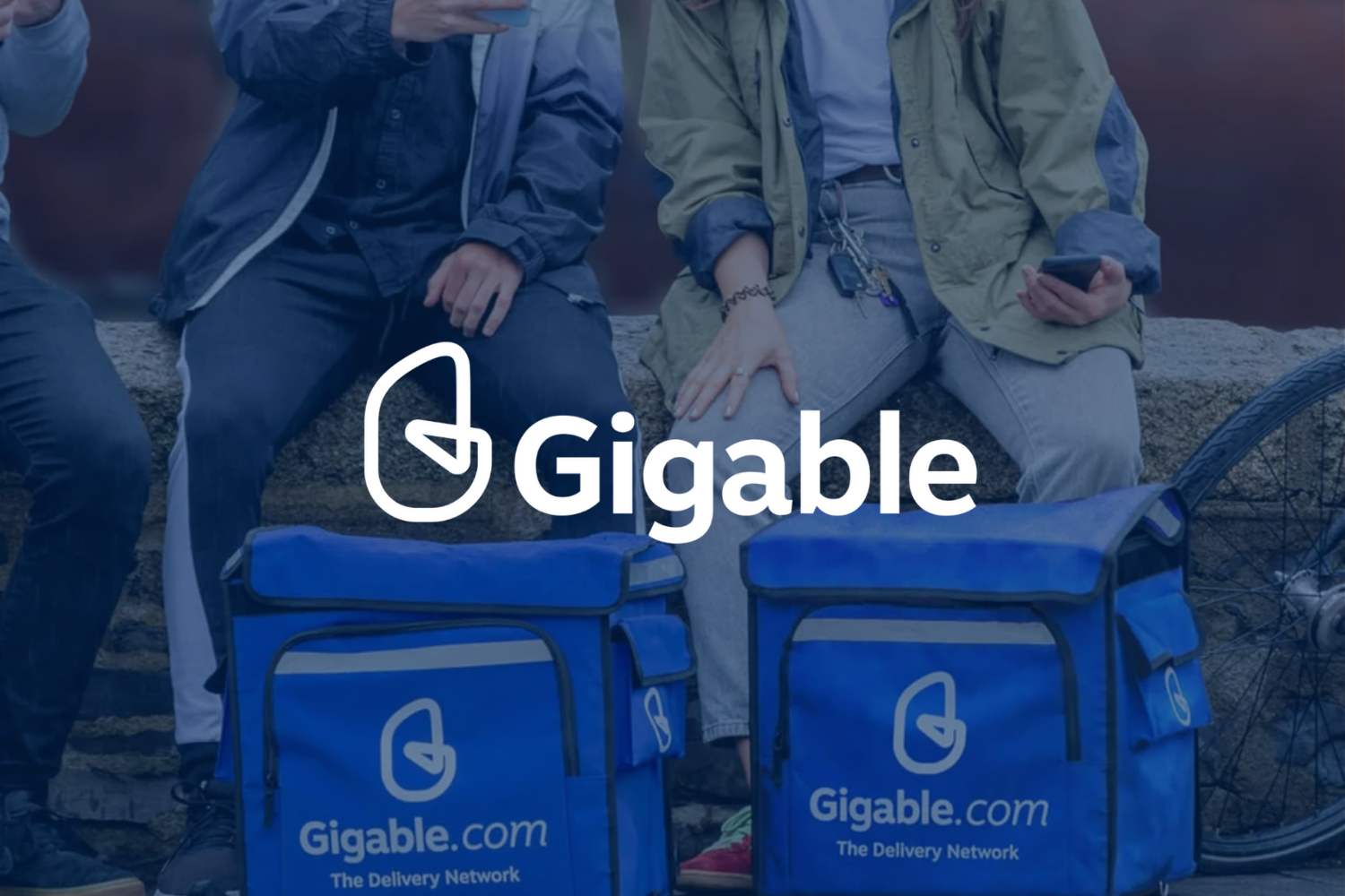 People sitting outdoors with Gigable branded delivery bags in front of them, some holding smartphones, near a bicycle and a stone wall.