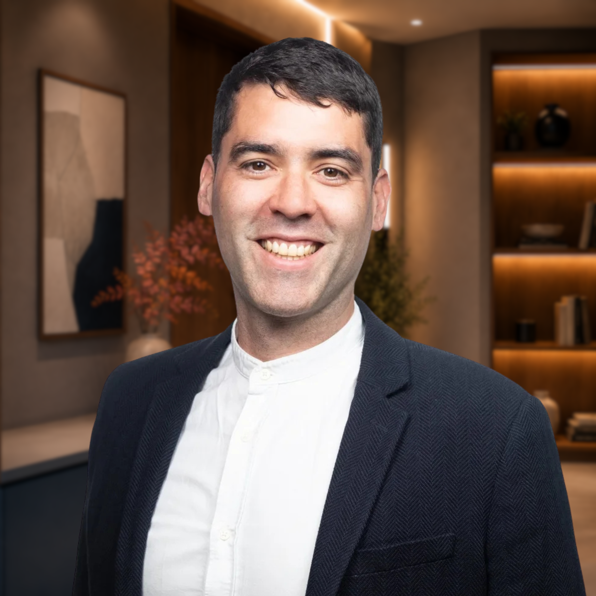 Close-up of a smiling man with dark hair wearing a white shirt and a dark blazer, in a warmly lit modern interior.
