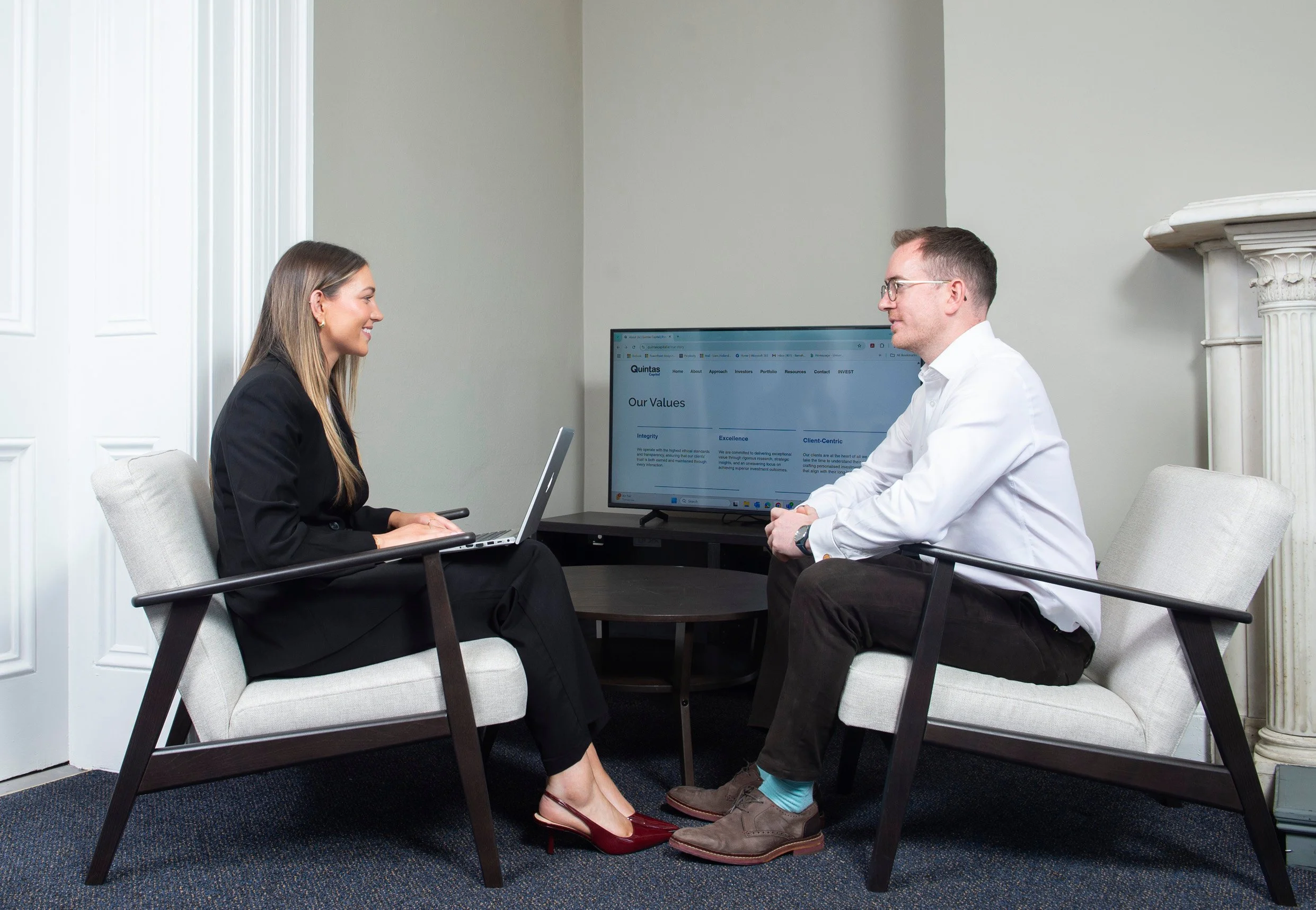 A man and woman in a professional office setting sitting across from each other. The woman is wearing a black blazer and red high heels, holding a laptop. The man is dressed in a white shirt and glasses. A monitor displaying a website about company values is on the table between them.