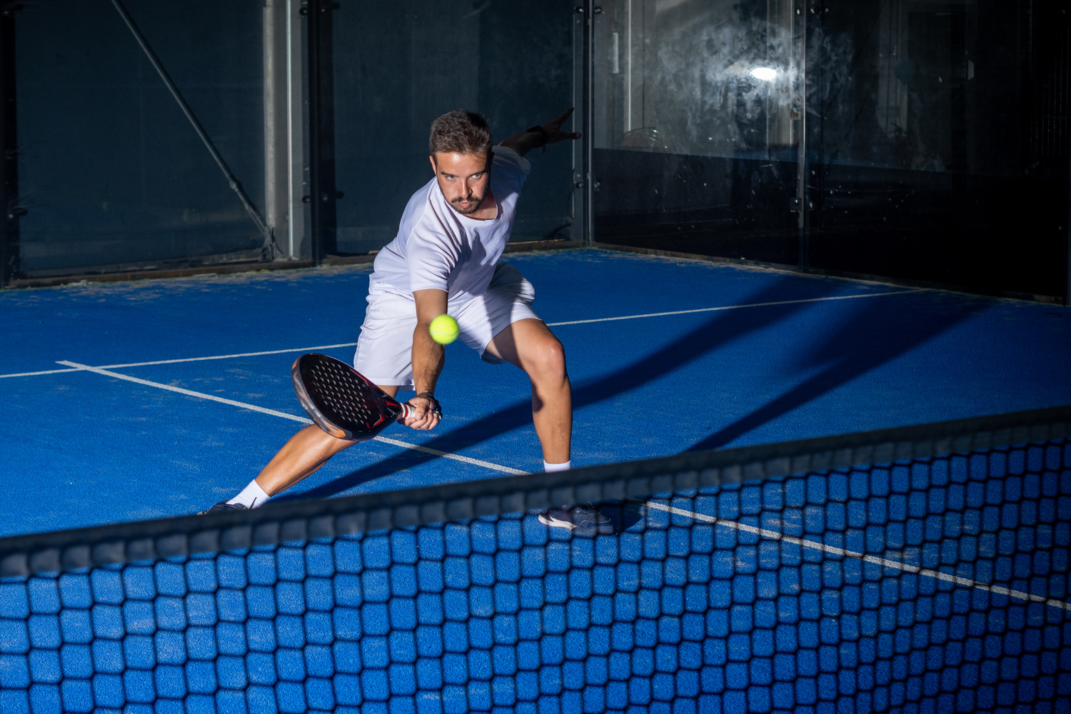 Male tennis player in white athletic clothing playing pickleball on blue indoor court, ready to hit the yellow tennis ball with paddle.