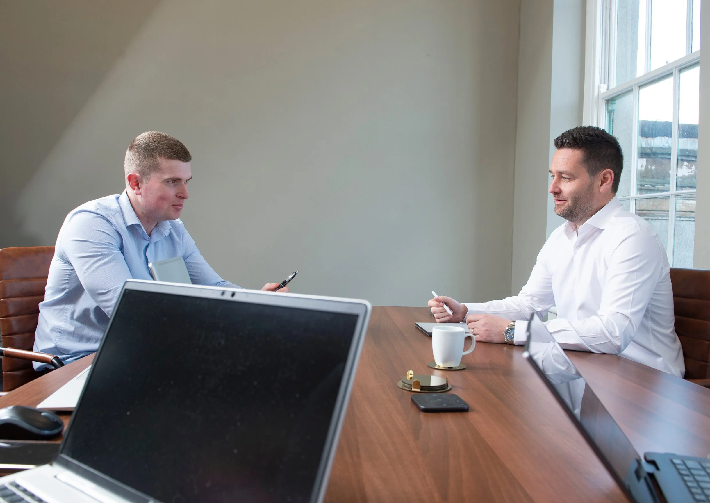 Two men in business attire sitting at a wooden conference table in a room with a large window. One man with short brown hair and a blue shirt is talking, and the other man with dark hair and a white shirt is listening. There are laptops, a coffee mug, a smartphone, and other office supplies on the table.