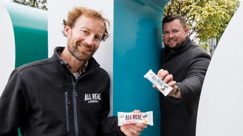 Two men smiling and holding protein bars labeled 'ALL REAL' outdoors.