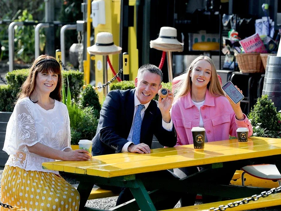 Three people sitting at a yellow outdoor table with drinks, smiling, holding a cupcake and a phone, with hats hanging on a stand behind them.