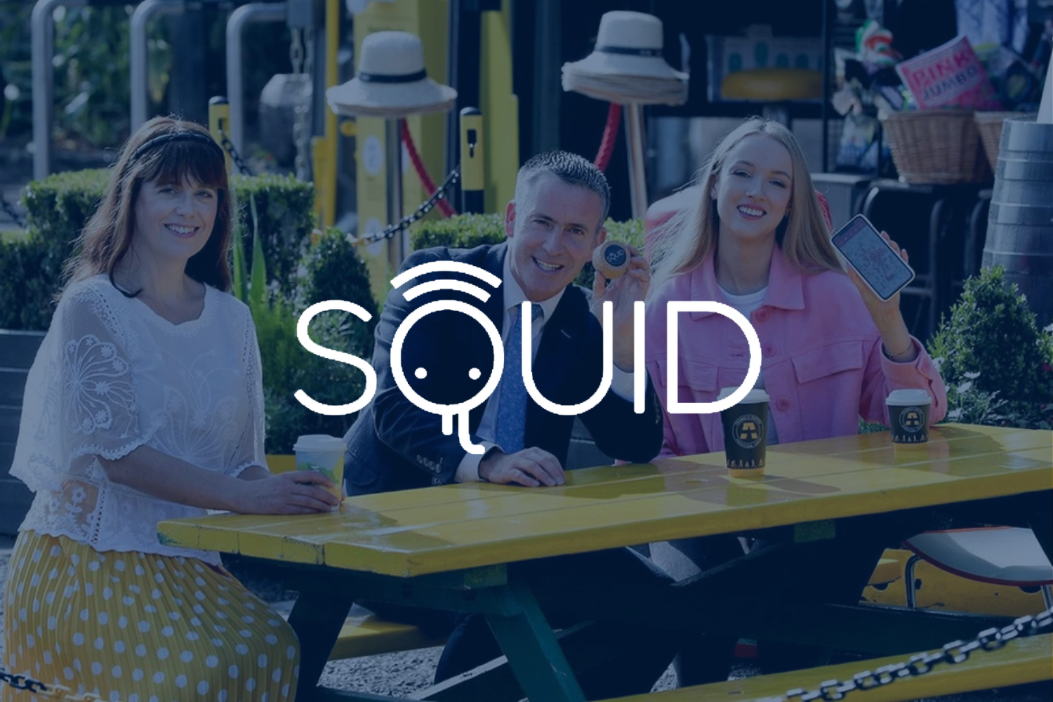 Three people sitting at a yellow picnic table outdoors, smiling and holding drinks and a phone, with a logo reading 'SQUiD' across the center.