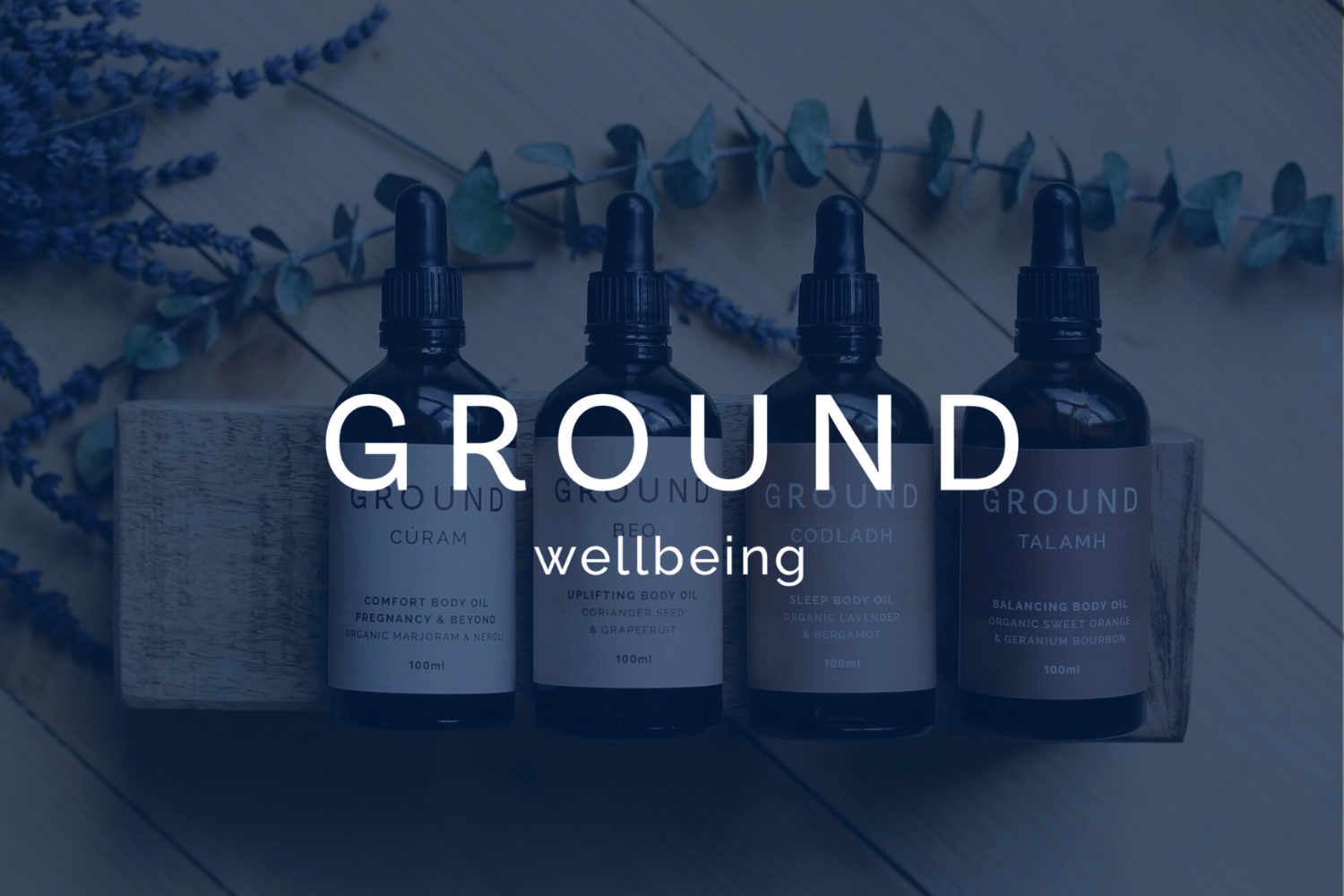 Four bottles of wellness body oils labeled ground with different scents, placed on a wooden tray, with dried lavender and eucalyptus in the background.