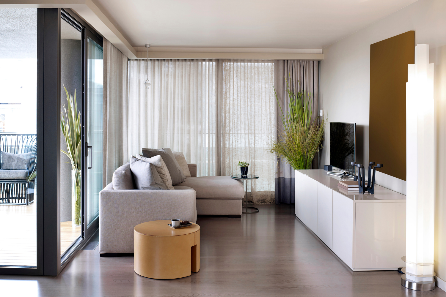 Modern living room with beige sofa, round yellow ottoman, flat-screen TV, white cabinet, large green plants, sheer curtains, and a balcony with outdoor seating.