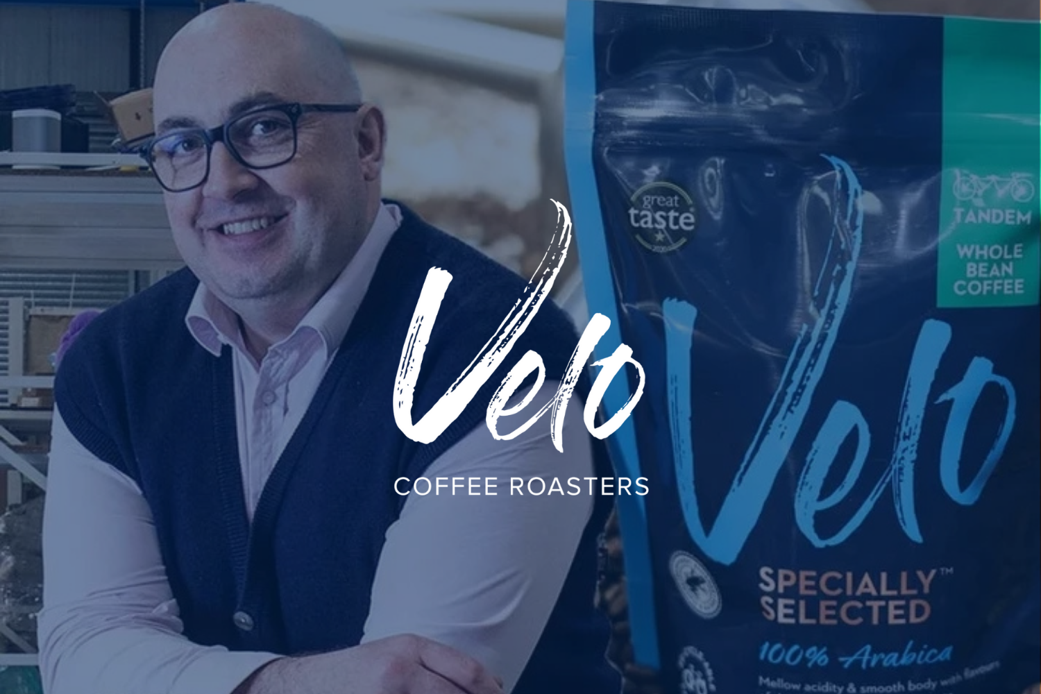 A man with glasses smiling, wearing a light-colored shirt and a dark vest, standing next to a package of Verbo Whole Bean Coffee in a store or warehouse setting.
