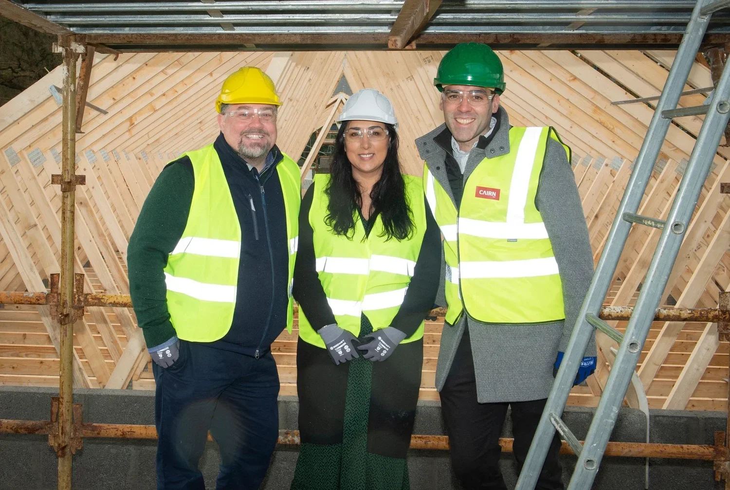 Three construction workers wearing safety vests and helmets standing inside a building under construction, with wooden framing and a metal ladder in the background.