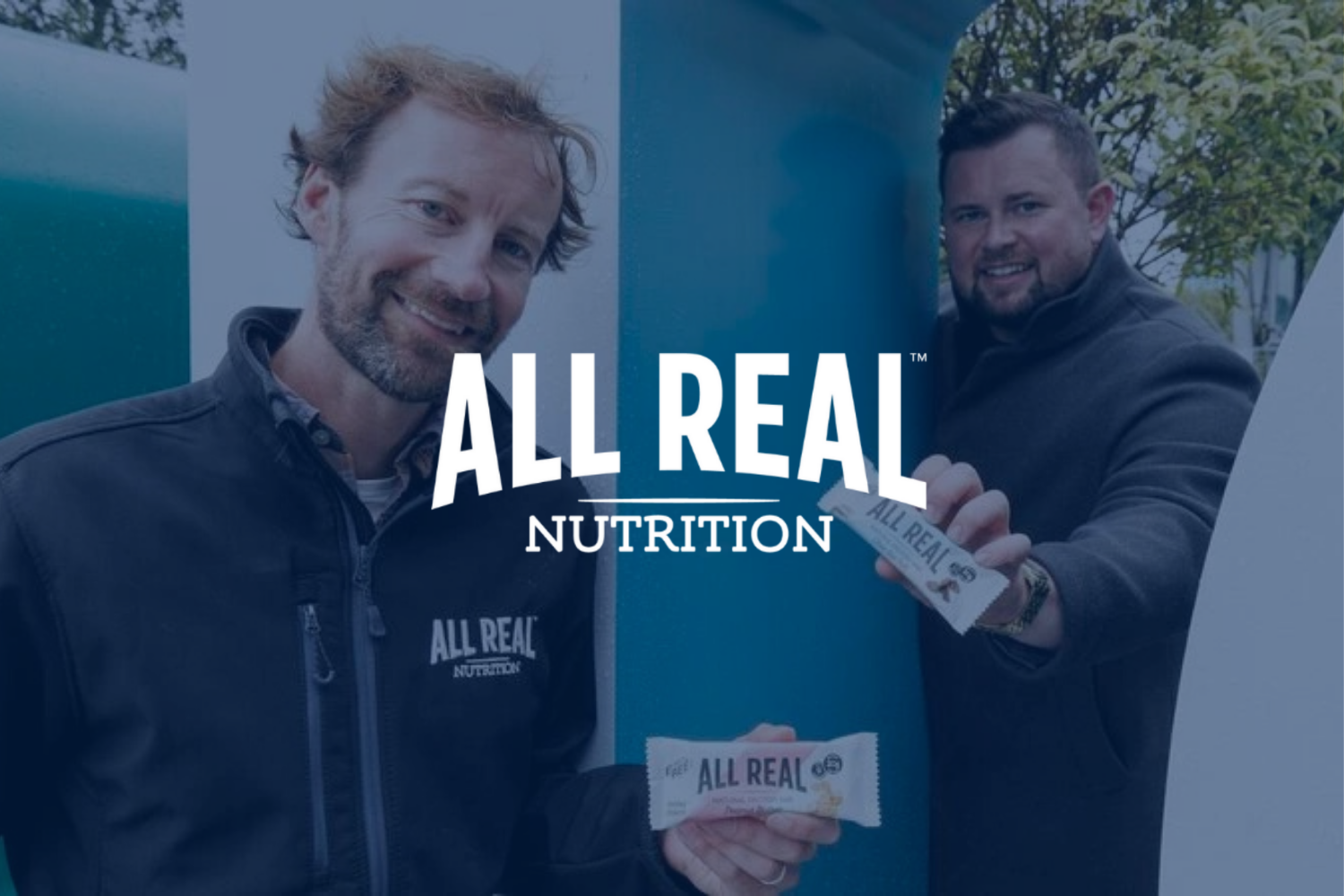 Two men smiling, one wearing a black jacket with 'ALL REAL NUTRITION' logo, holding a snack bar; the other in a dark jacket, also holding a snack bar. They are outdoors near a blue wall and trees, promoting All Real Nutrition.