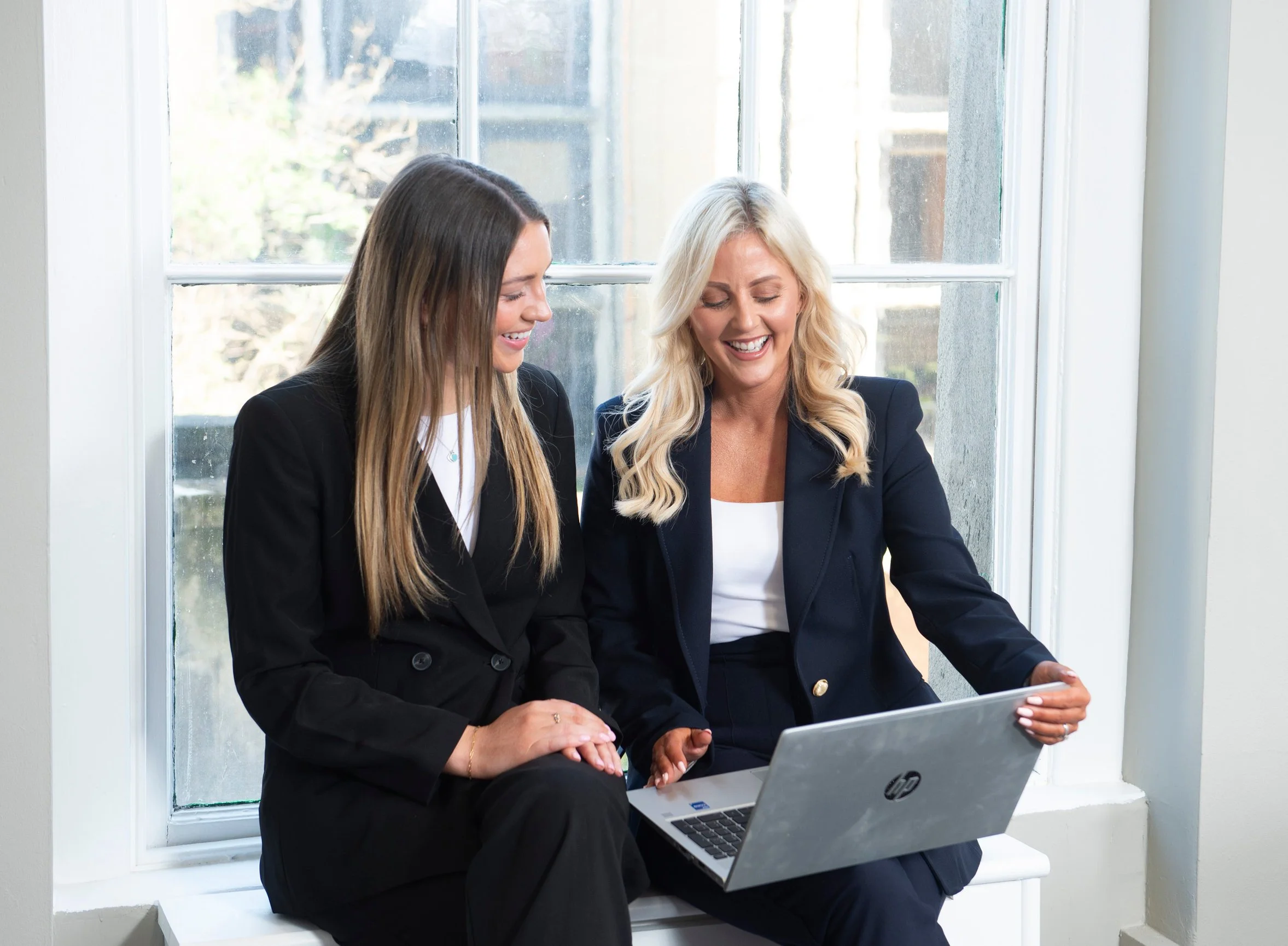 Two women in business attire sitting by a window looking at a laptop and smiling.