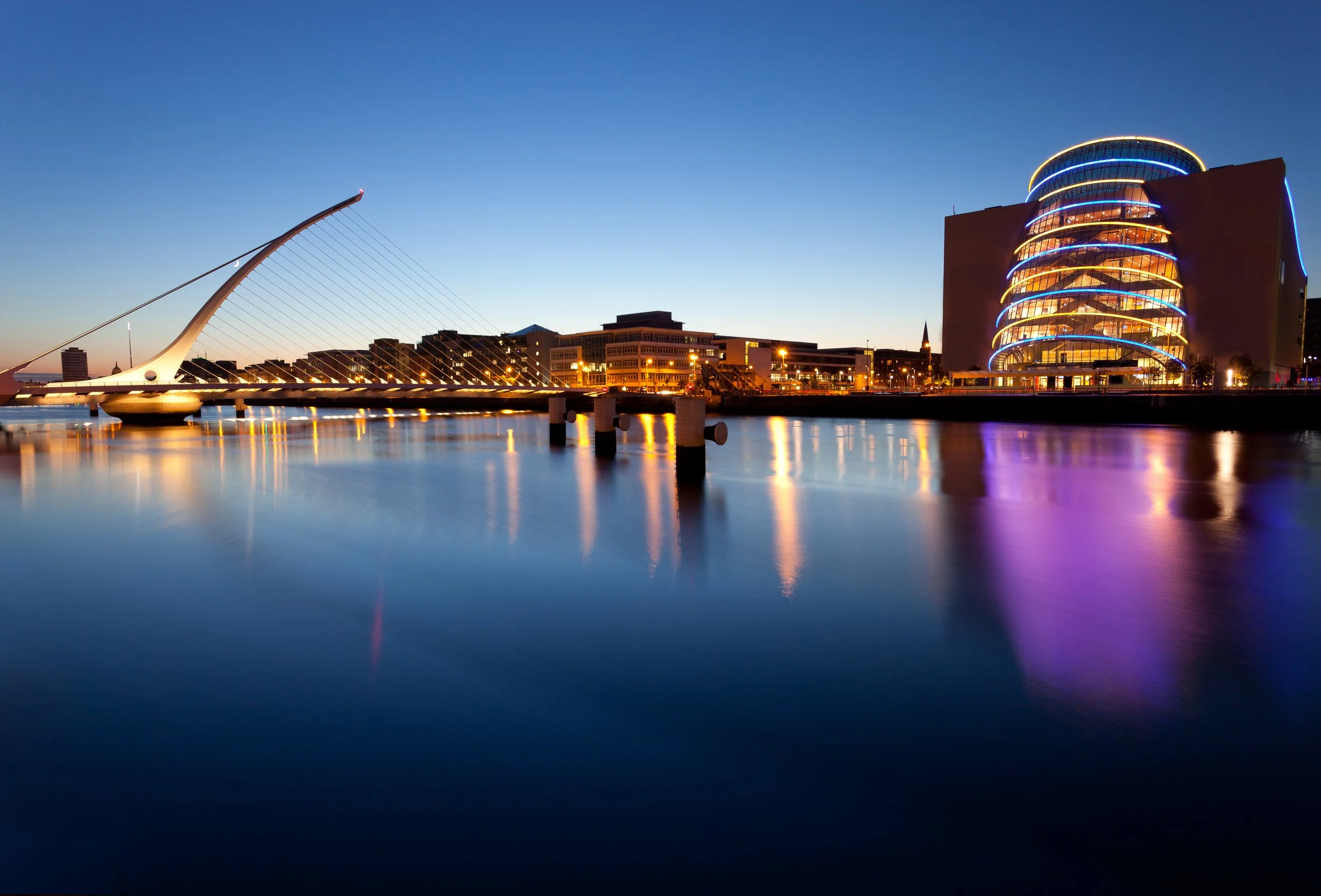 Nighttime cityscape featuring a modern building with curved, illuminated blue and yellow lines, a bridge with a unique cable design, and water reflecting the city lights.
