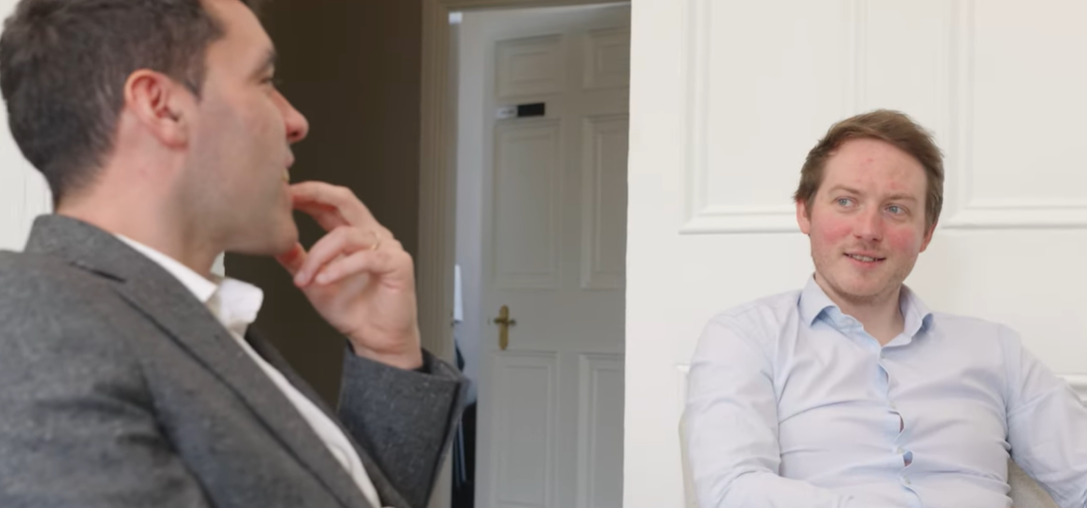 Two men are sitting and talking in a white room, one in a suit and the other in a light blue shirt, with a door in the background.