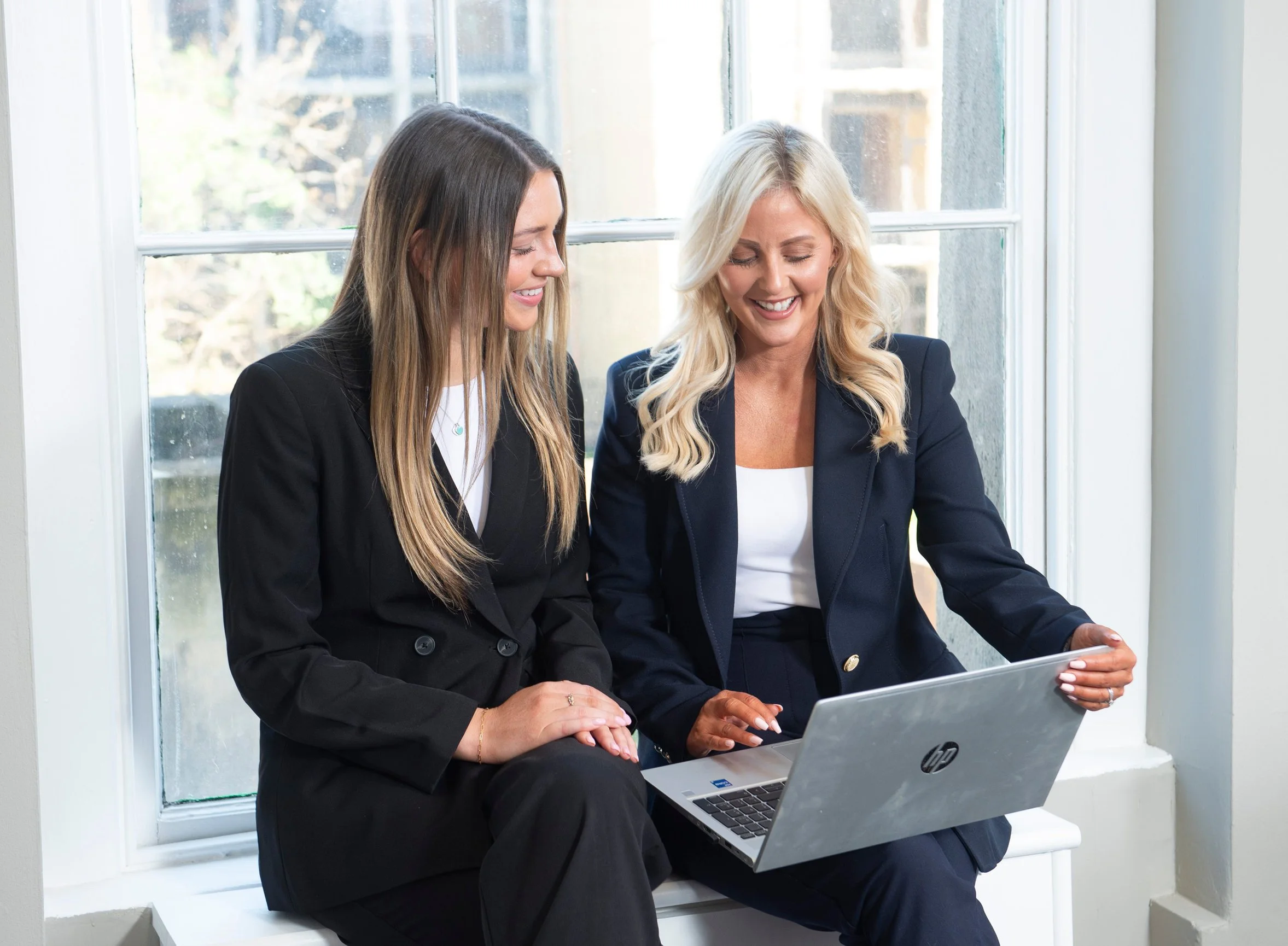 Two women in business attire sitting on a windowsill looking at a laptop, smiling.