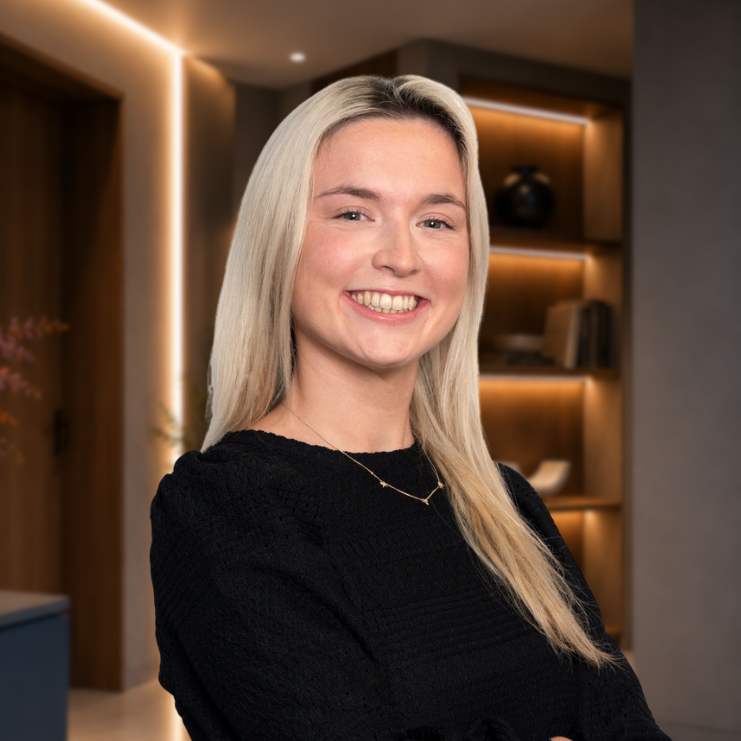 Close-up of a young woman with long blonde hair smiling in an indoor setting with warm lighting and modern decor.