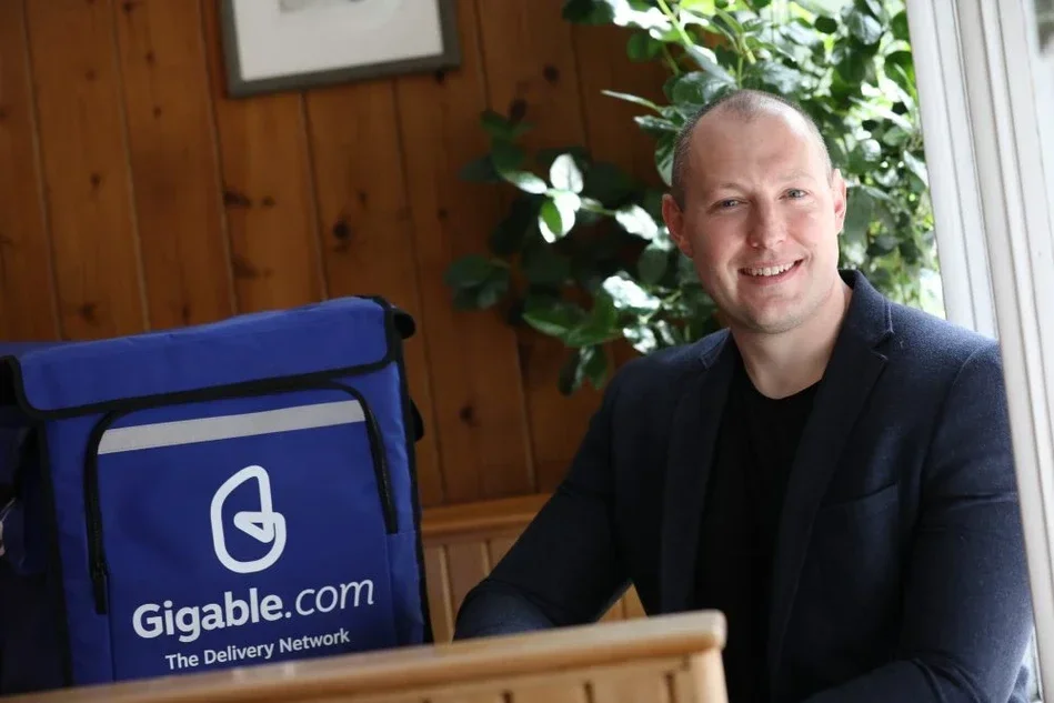 A smiling man in a dark blazer sitting at a wooden table near a window with green plants in the background, next to a blue Gigable.com delivery bag.