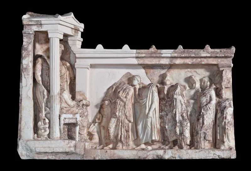 Name: Relief in the form of a chapel and a stoa.
Creator: N/A.
Dating Back To: Around 350 BC.
Material: Penteli Marble.
Found In: 1938 in the sanctuary of Asclepius.