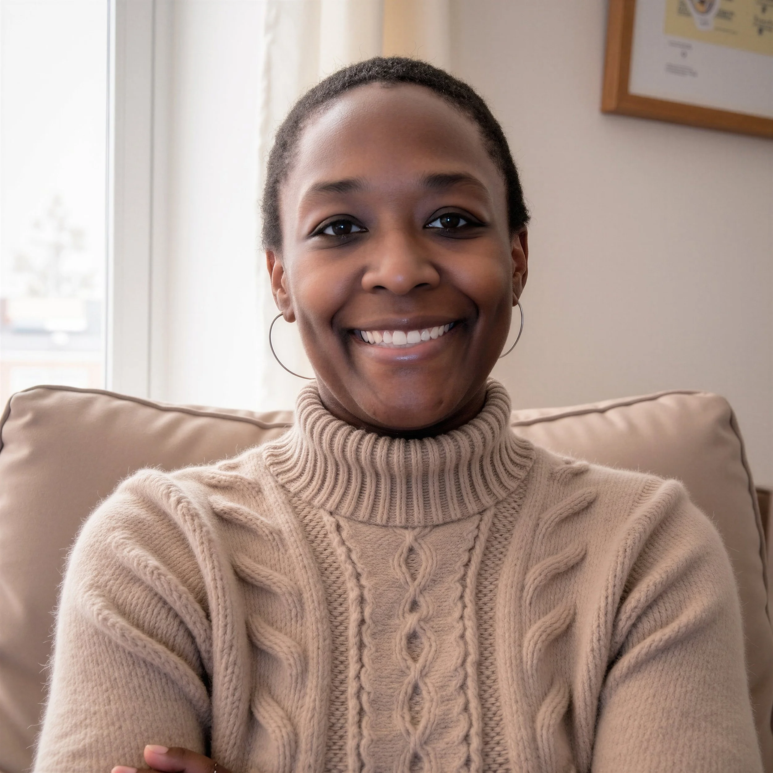 Close-up of a smiling woman with short hair wearing a beige turtleneck sweater, sitting indoors near a window with a wooden frame and a wall displaying a framed certificate.