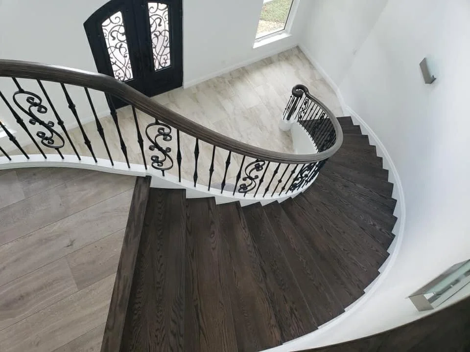 A spiral staircase with dark wooden steps, a black ornate metal railing, and a white wall with a large window and a decorative front door with ironwork by Hamilton Stair and Rail.