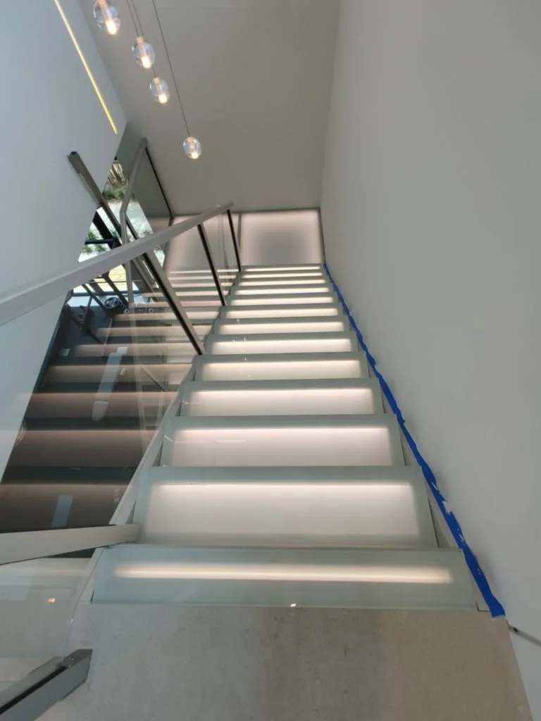 Modern staircase with illuminated glass steps, black metal railing, glass wall on the left, hanging pendant lights, and a large window at the top, in a sleek interior space by Hamilton Stair and Rail.