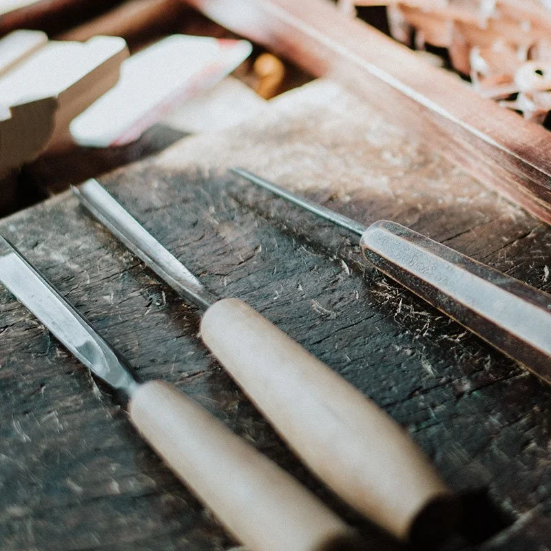 Three woodworking carving tools with wooden handles resting on a dark, weathered wooden workbench.