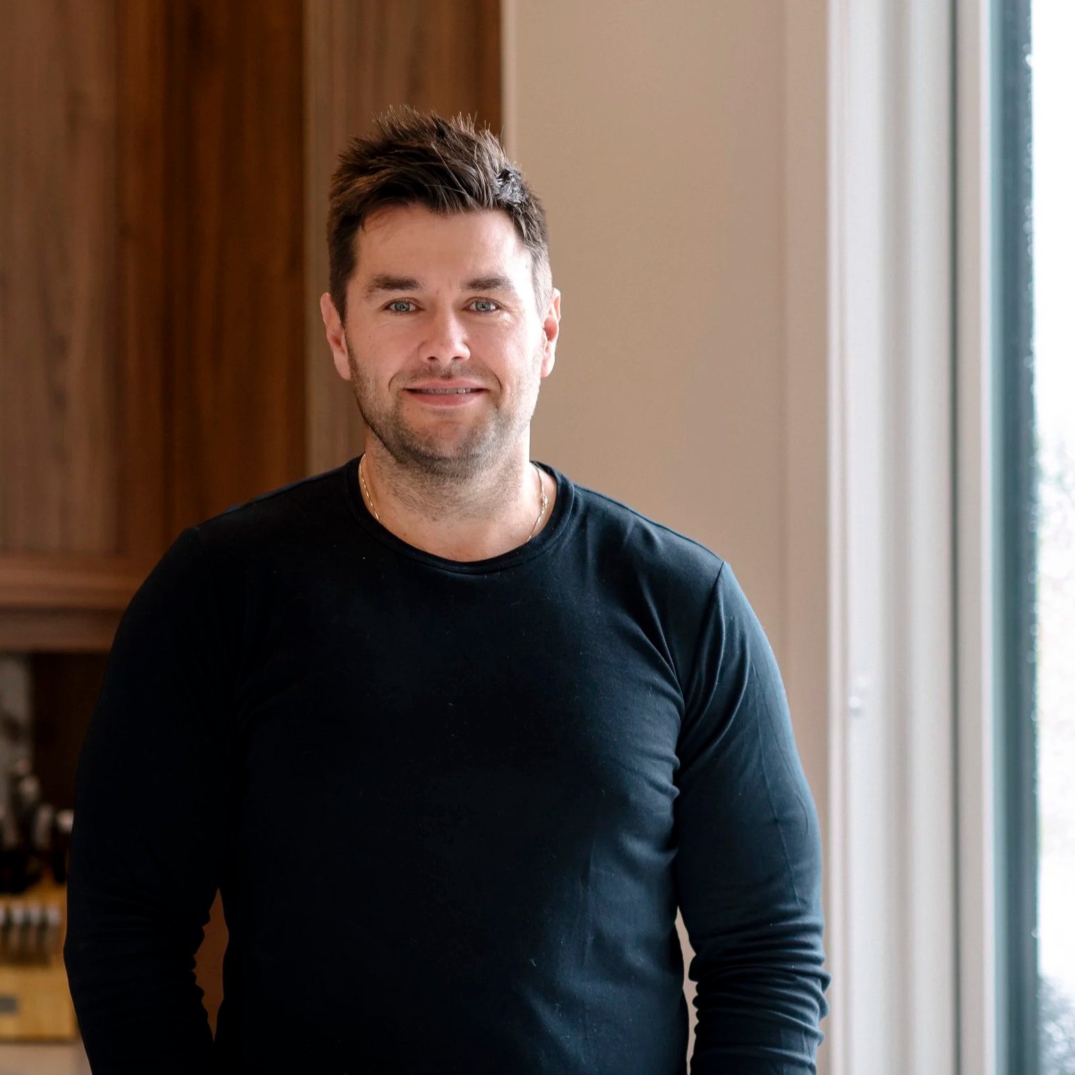 A young man with dark hair and a beard, wearing a black long-sleeve shirt, standing indoors near a window with curtains, smiling at the camera.