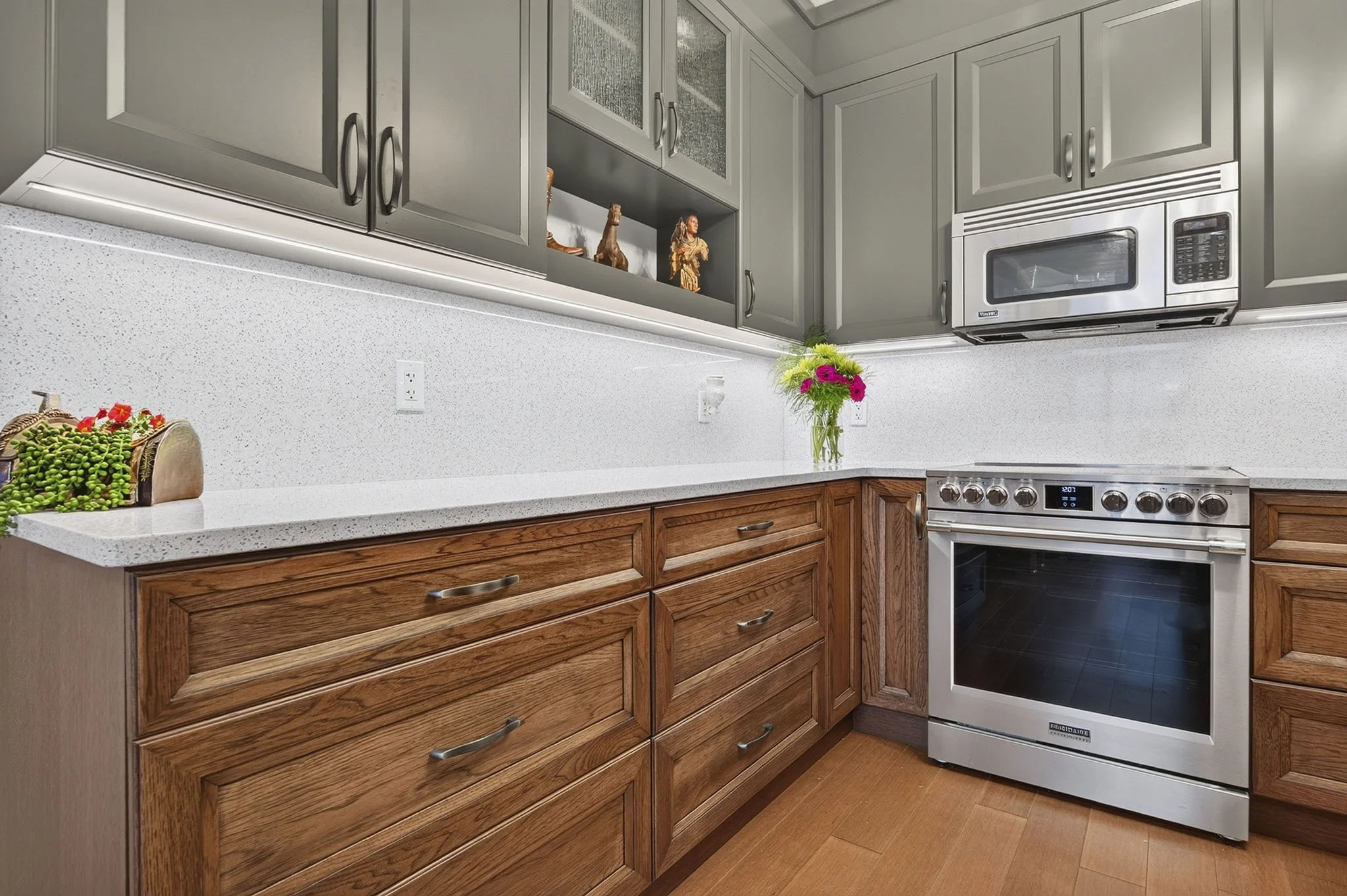 A kitchen with wooden cabinets, white speckled countertops, and stainless steel appliances, including a microwave and oven. There is a vase with pink and green flowers on the counter and decorative items on an open shelf above.
