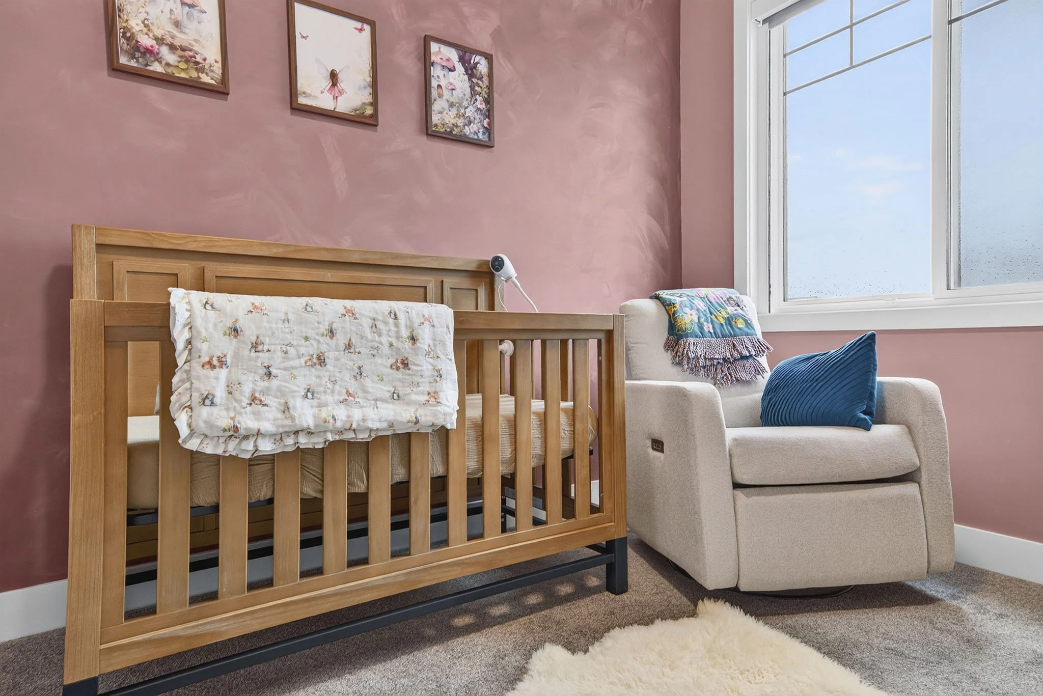 A cozy bedroom with pink textured walls, decorated with three framed pictures of fairies, flowers, and butterflies. There is a wooden crib with a mobile and a white blanket featuring colorful animal prints. Next to the crib, a beige armchair with a m