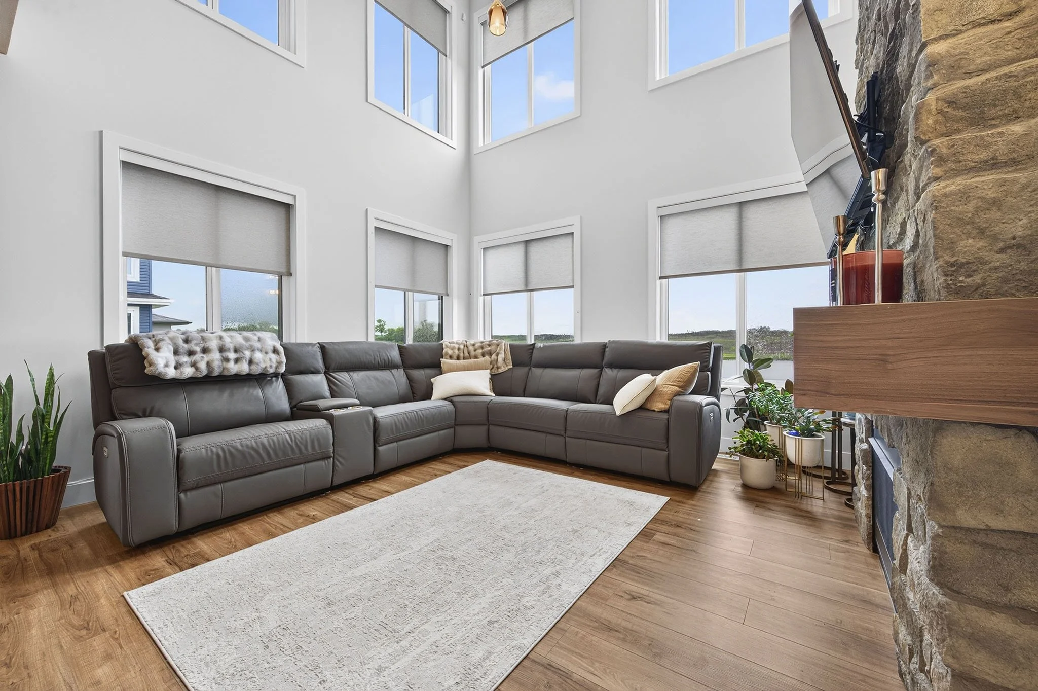 Living room with a large sectional sofa, multiple windows with white blinds, and a fireplace with a stone wall and a wooden mantel. Potted plants are on the floor, and there is a ceiling light fixture.
