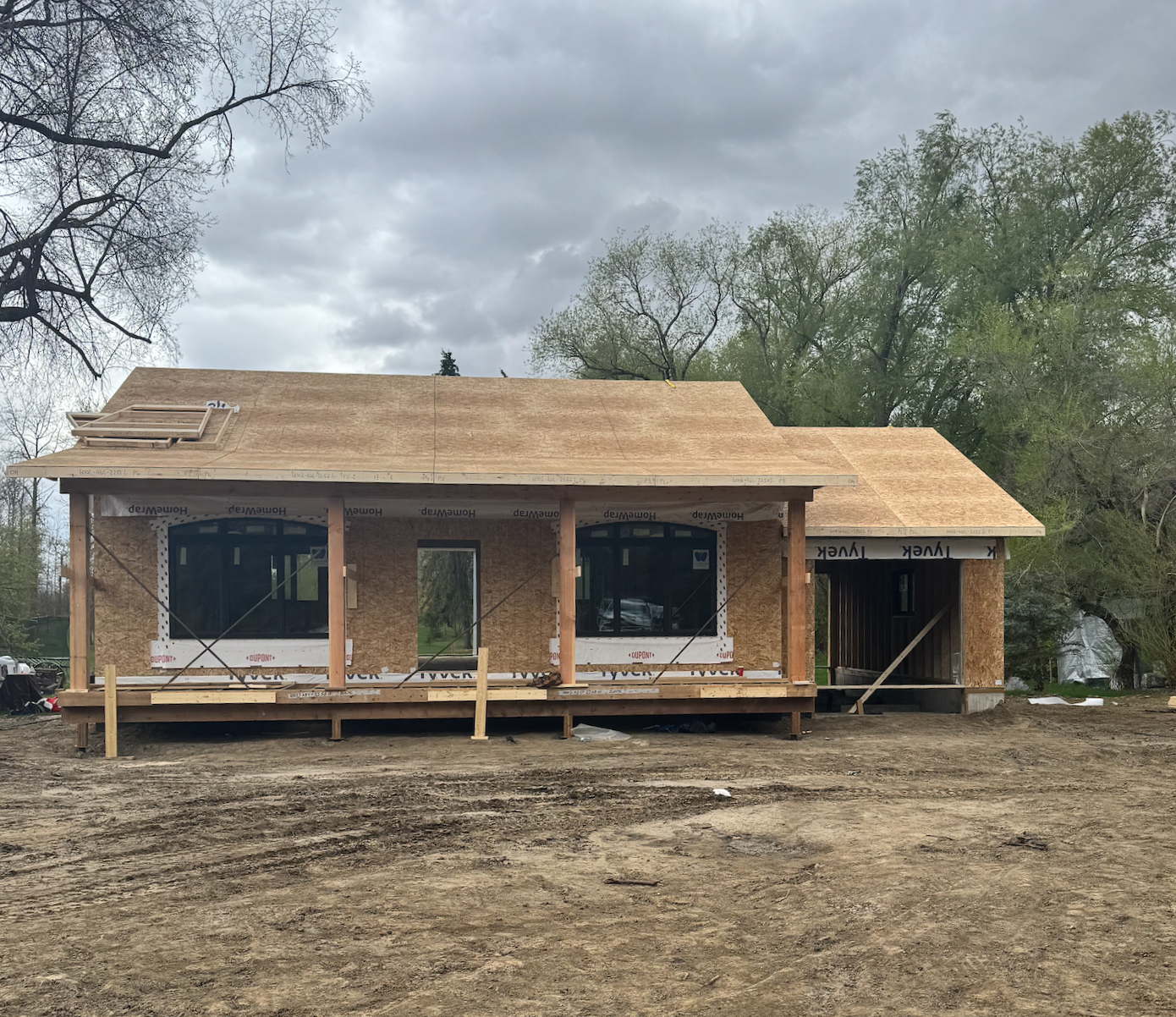 A garden suite under construction with wooden framing and plywood sheathing, windows installed, and a prepared roof, set outdoors with trees and cloudy sky in the background. Copyright CJ Living Co.