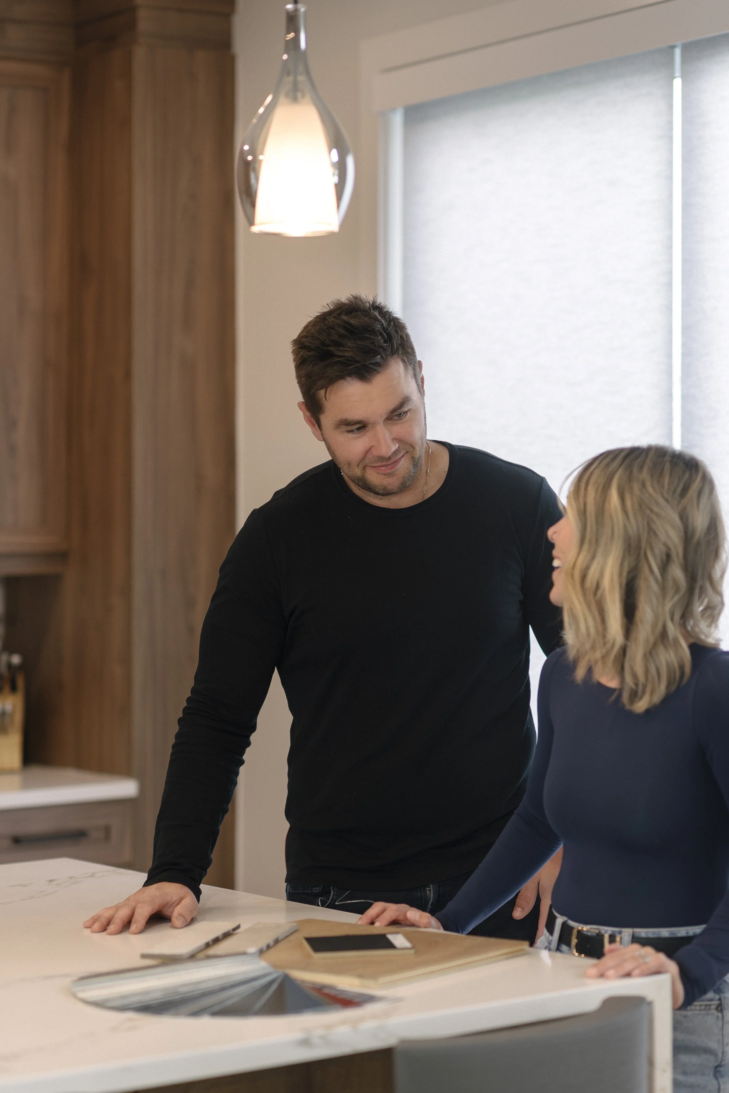 A man and a woman are engaged in a conversation or discussion while leaning on a table in a modern room with wood paneling and a large window.