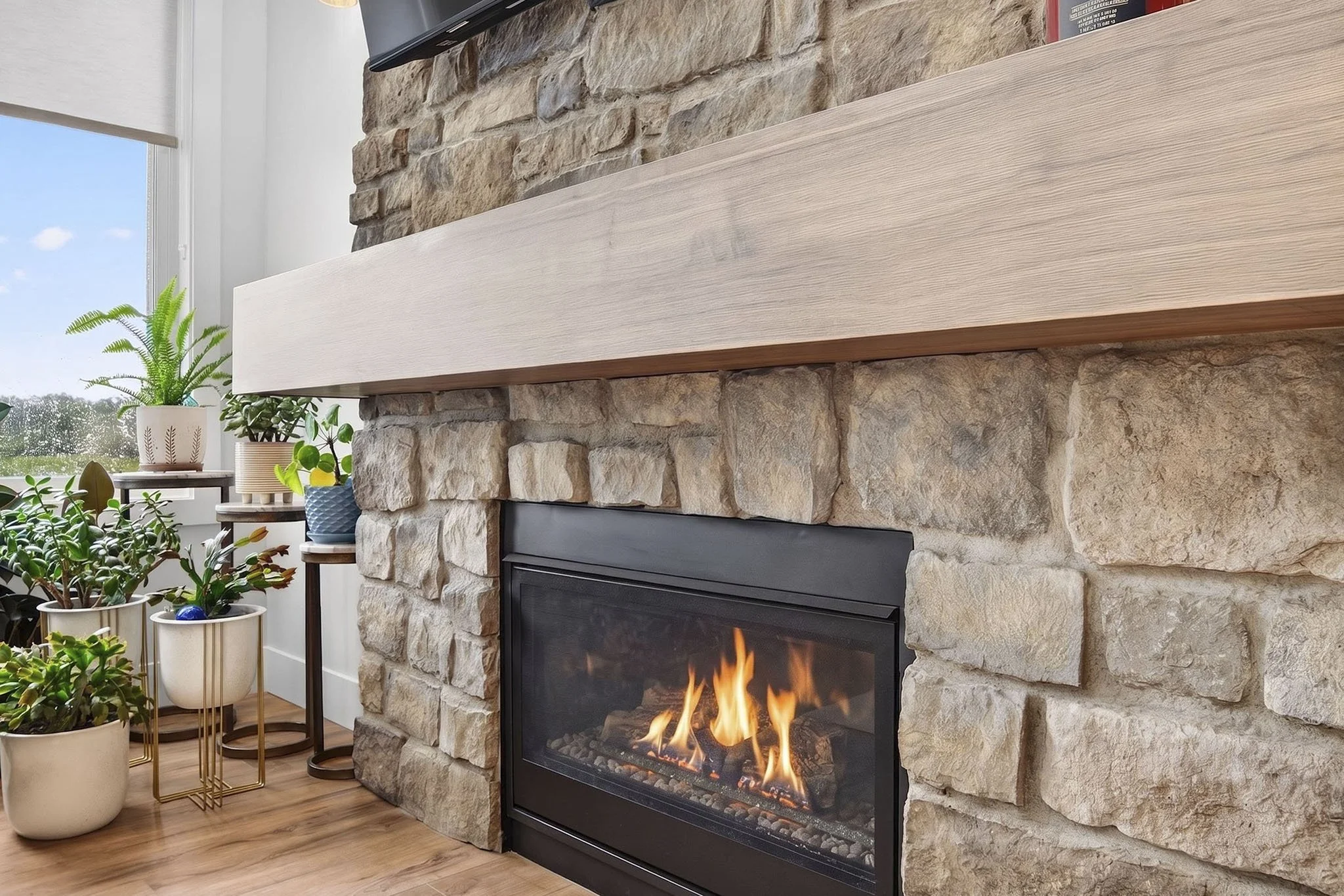 Living room with a stone fireplace, wooden mantel, and potted plants near a window showing a cloudy sky.
