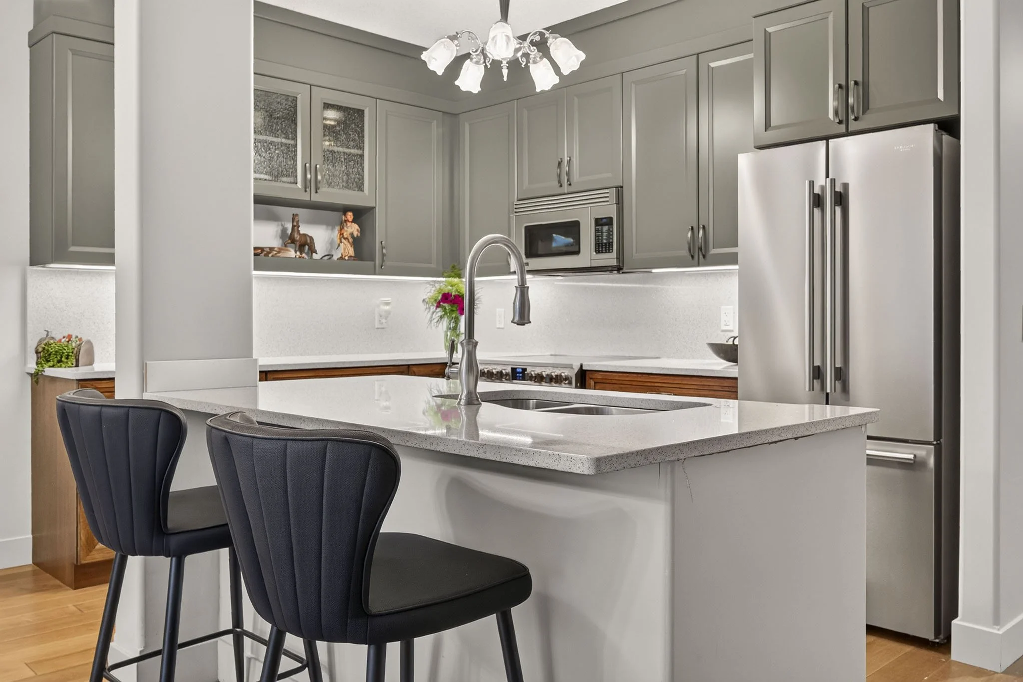 Modern kitchen with gray cabinets, white countertop island, stainless steel refrigerator, microwave, and chandelier. Two black chairs at the island and a small vase of pink flowers.