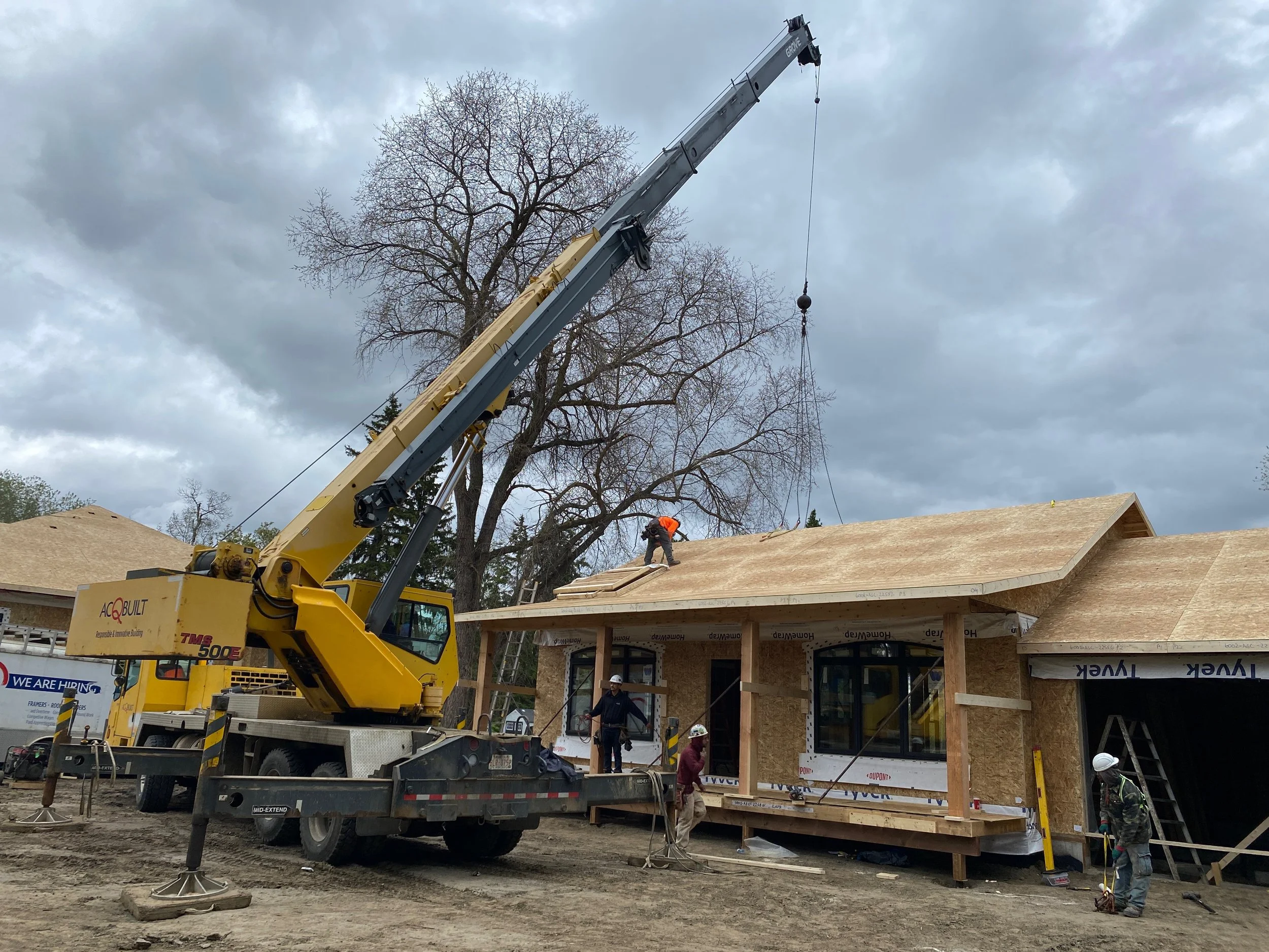 Construction workers building a garden suite with wooden framework, using a large crane to lift materials, under a cloudy sky. Copyright CJ Living Co.