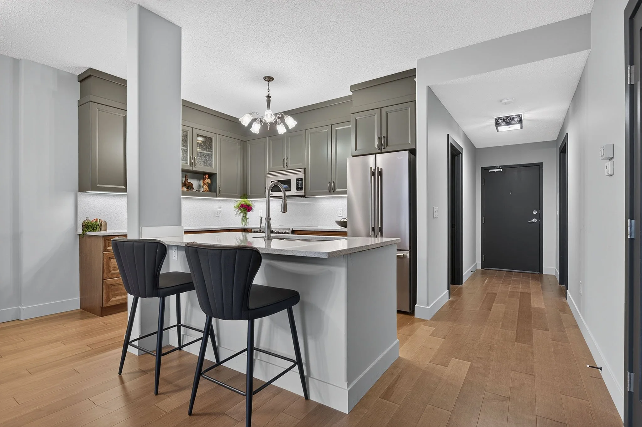 A modern kitchen with gray cabinets, a white island with a sink, black chairs, a stainless steel refrigerator, a microwave, decorative figures, a flower vase, and a hallway with wood flooring and a black front door.