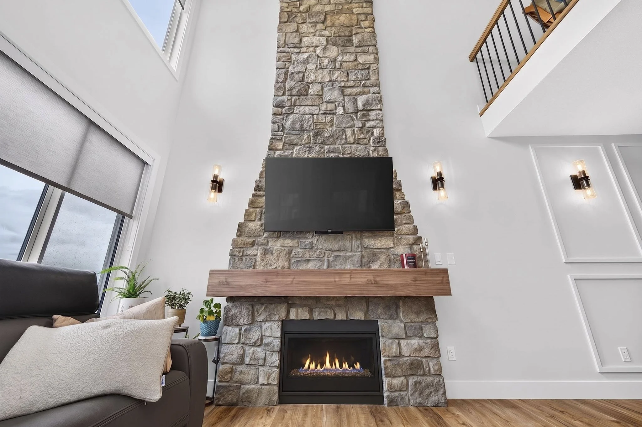 Living room with a stone fireplace, mounted flat-screen TV, and modern wall sconces. Large window with a roller shade, ceiling with loft railing, and hardwood floor.