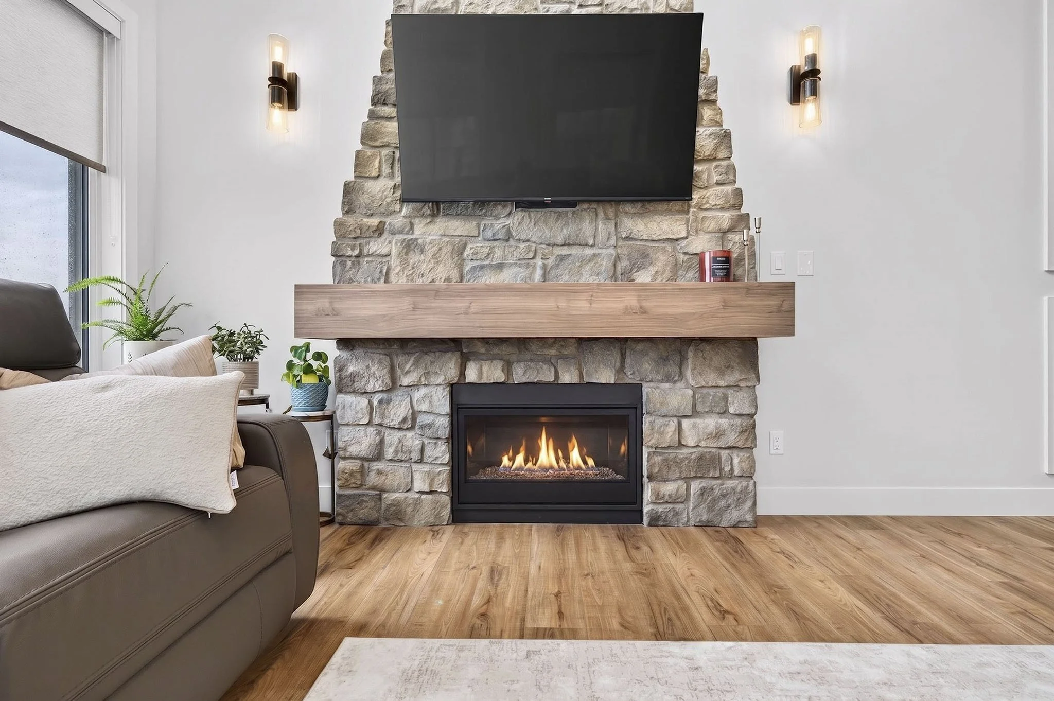 Living room with a stone fireplace topped with a wooden mantel, a mounted flat-screen TV above it, and a cozy fire burning inside. Comfort items include a sofa, a potted plant, and a rug, with wooden flooring and wall-mounted lighting fixtures.