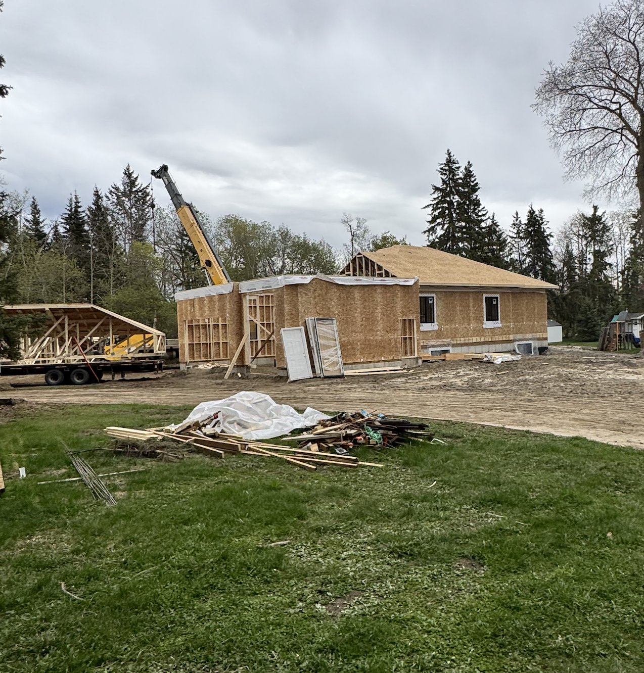 House under construction with wood framing, a crane, construction material, and a partially finished roof on a cloudy day.