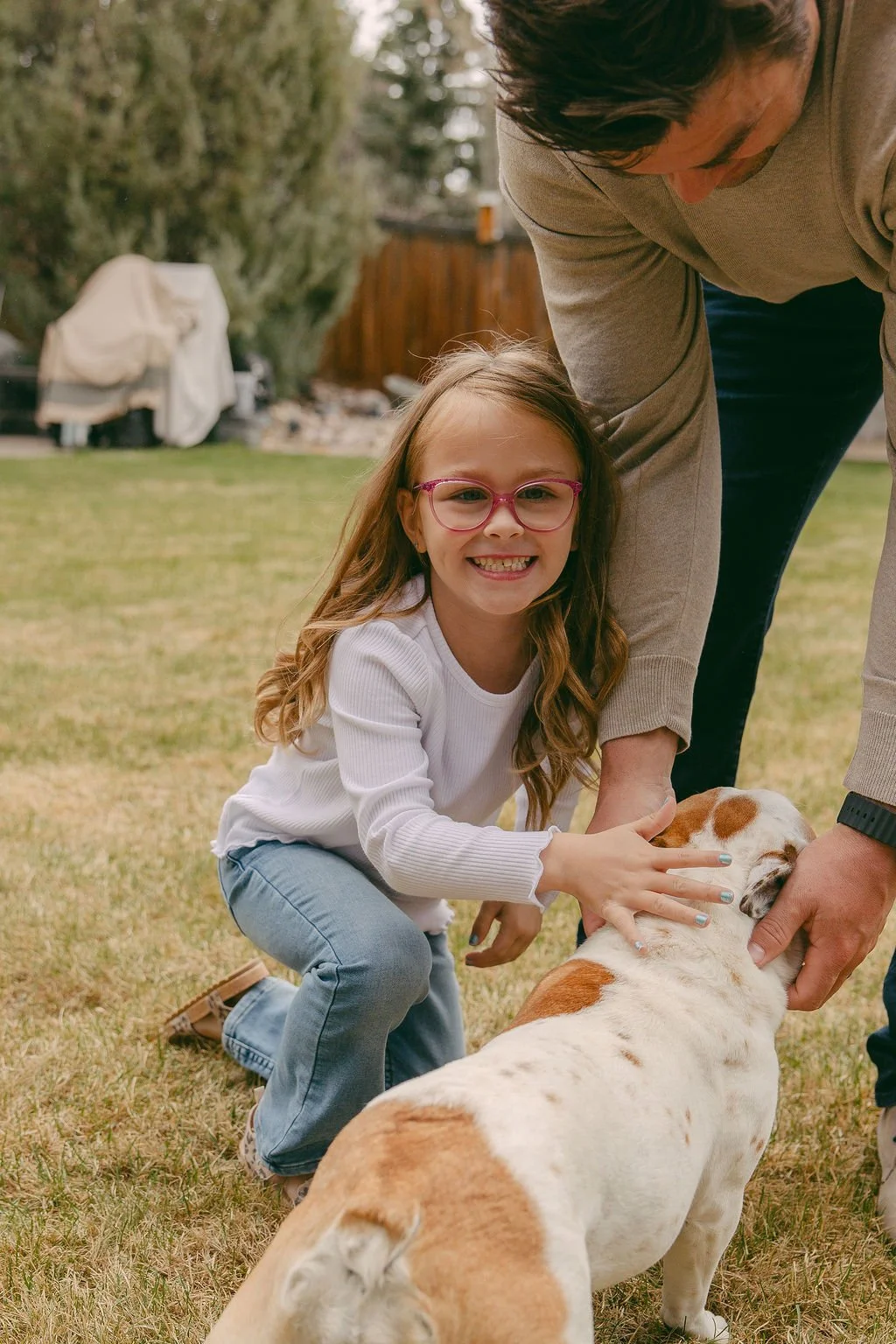 Blake Foster, cancer survivor, with glasses, smiling, kneeling on grass while petting a white and brown dog outside. An adult is helping her hold the dog's head. Trees and a wooden fence are in the background.