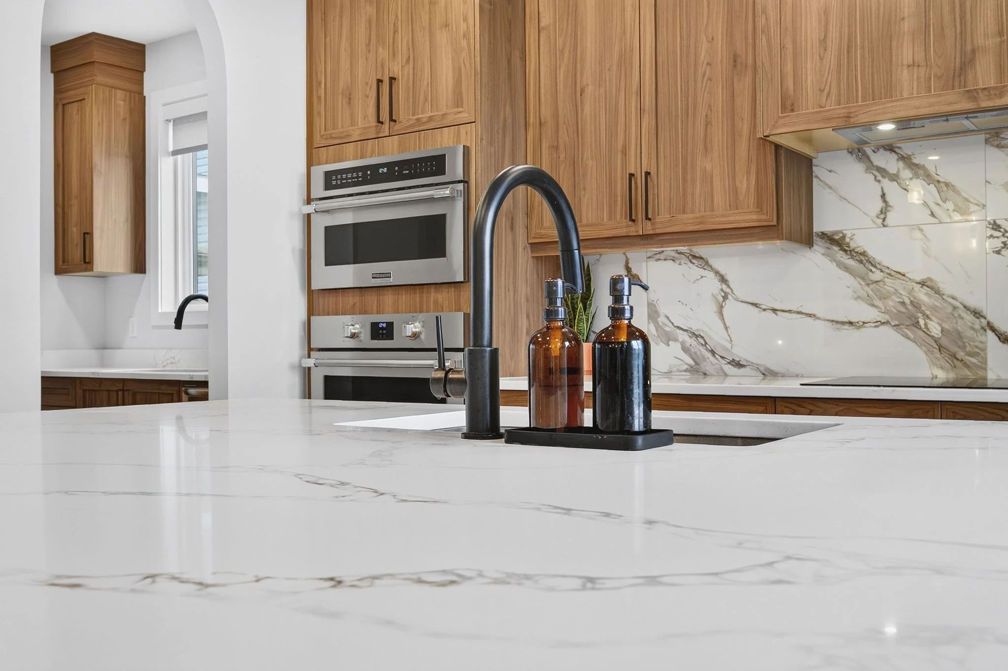 Modern kitchen with white marble countertop and backsplash, wooden cabinets, black sink faucet, and soap dispensers.