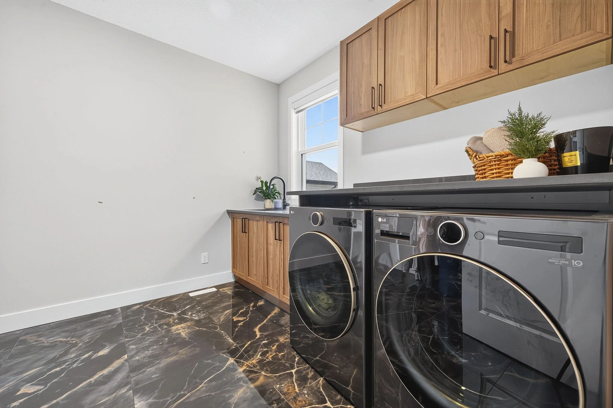 A laundry room with black marble floors, wooden cabinets, a washing machine, a basket with a plant, and a window.